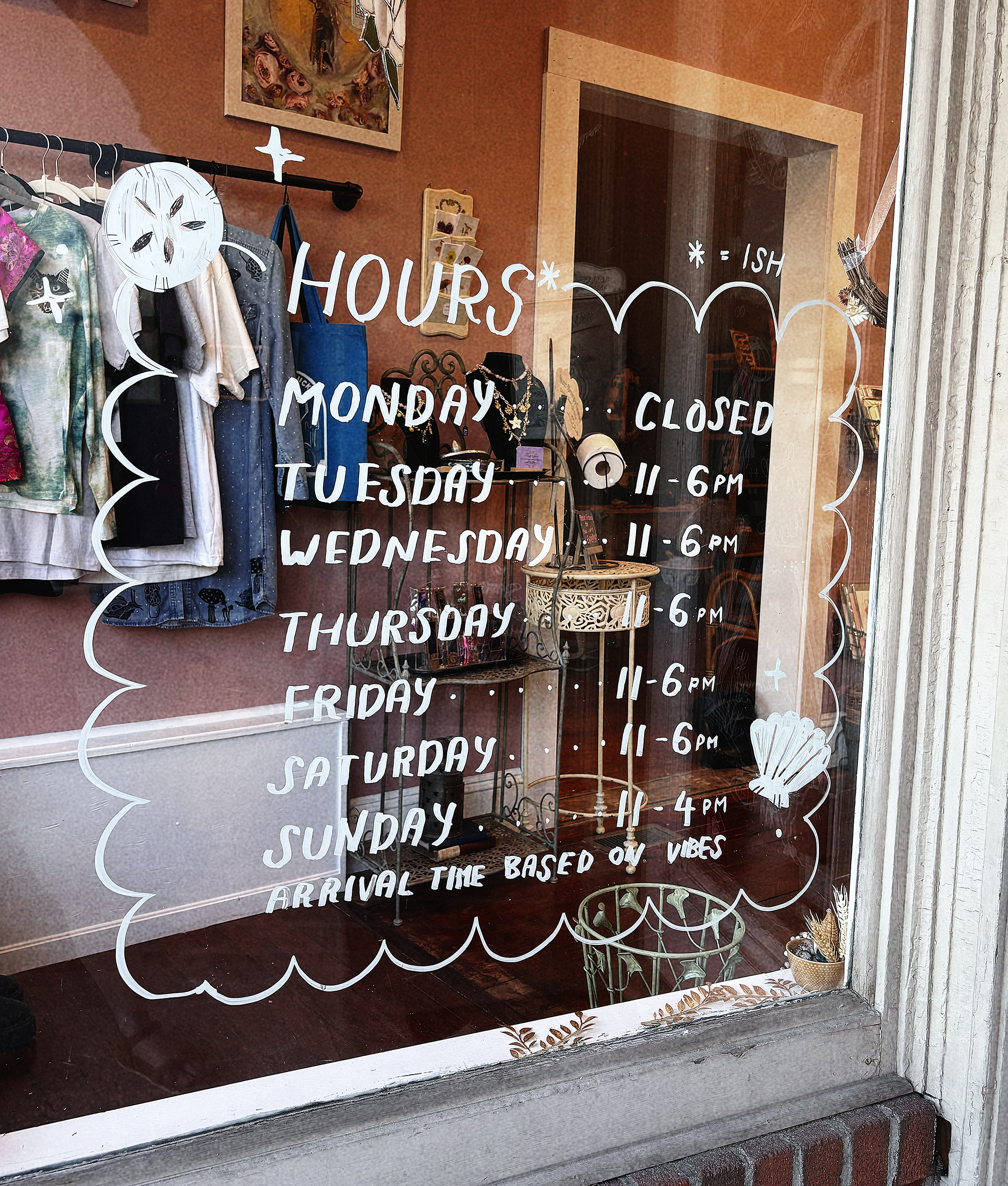 A store window with white hand-painted store hours from 11 a.m. to 6 p.m. and shell decorative accents.