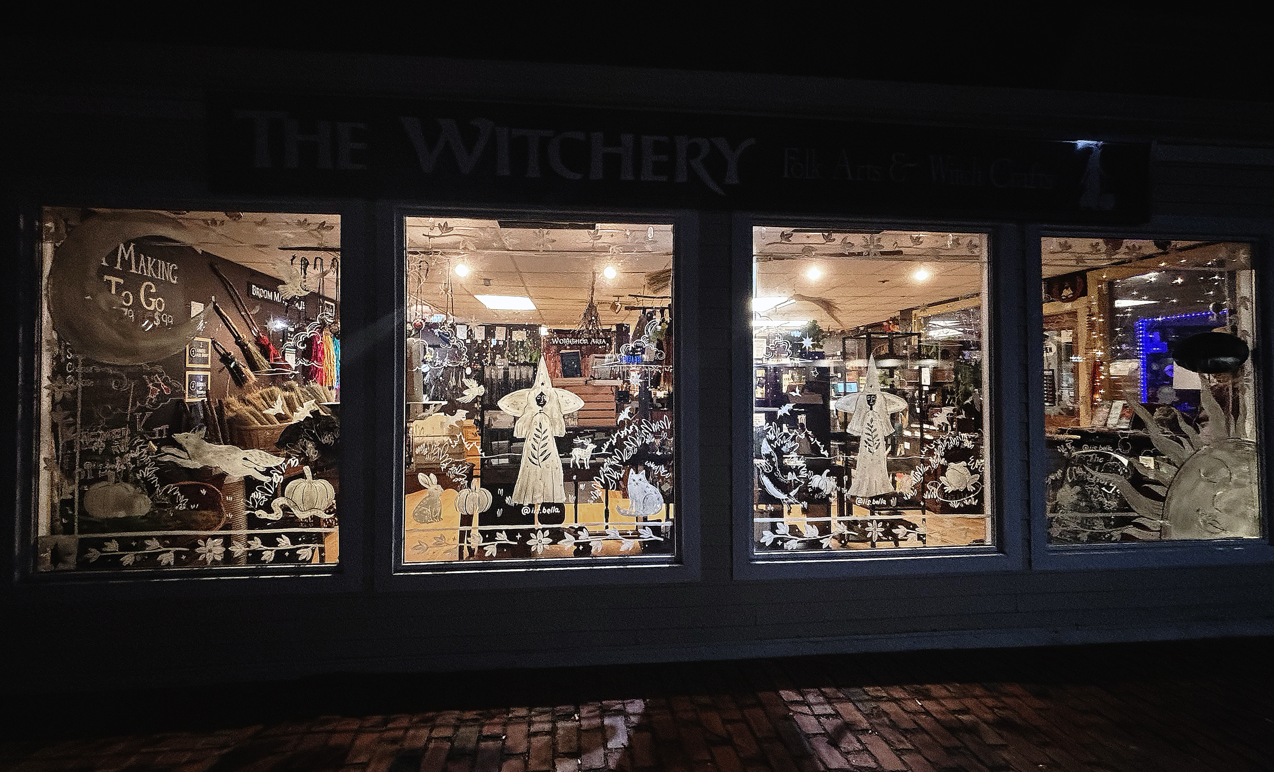 Hand-painted storefront window mural with autumn and Halloween-themed artwork, featuring pumpkins, witches, and animals.