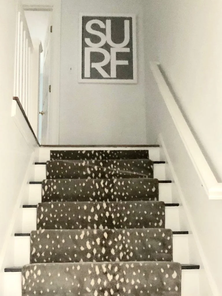 “When I started the project, the first thing that hit me was the staircase,” says Jerde. “It’s right in the middle of the house, but the rest of the house is an open floor plan. I needed to treat these stairs special, otherwise they’re an intrusion.” Jerde says they decided to wrap the stairs in white shiplap. “It made quite a difference to the whole house.” The staircase boasts an antelope-print runner, and hanging on the wall at the top of the stairs is a complementing art piece reading SURF, from Bodega Nantucket.