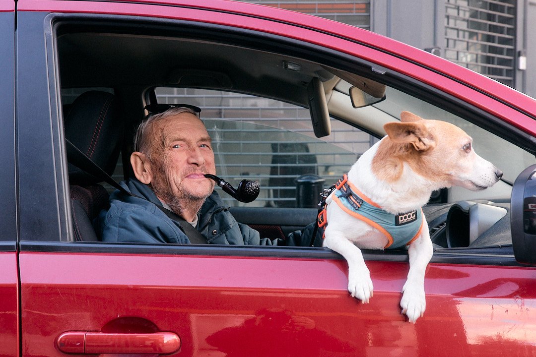 Street portrait in London of man smoking pipe in red car smiling with jack russell dog leaning out window