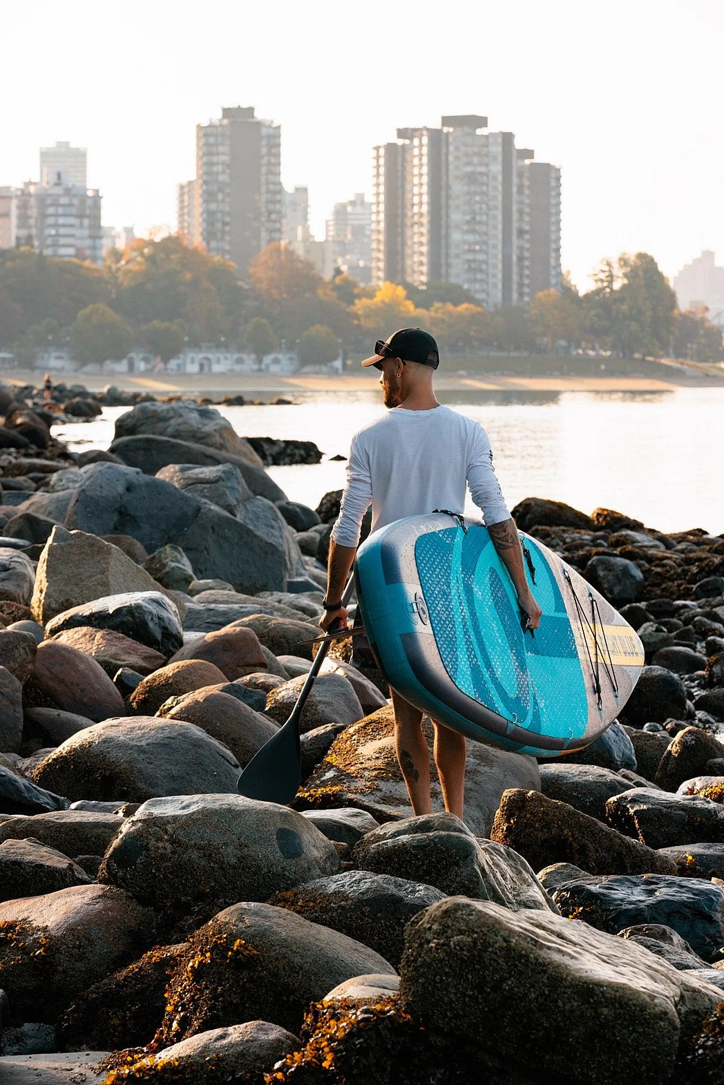 Dating photography portrait of man with cap walking on rocky shore holding paddle board with building skyline and water in background