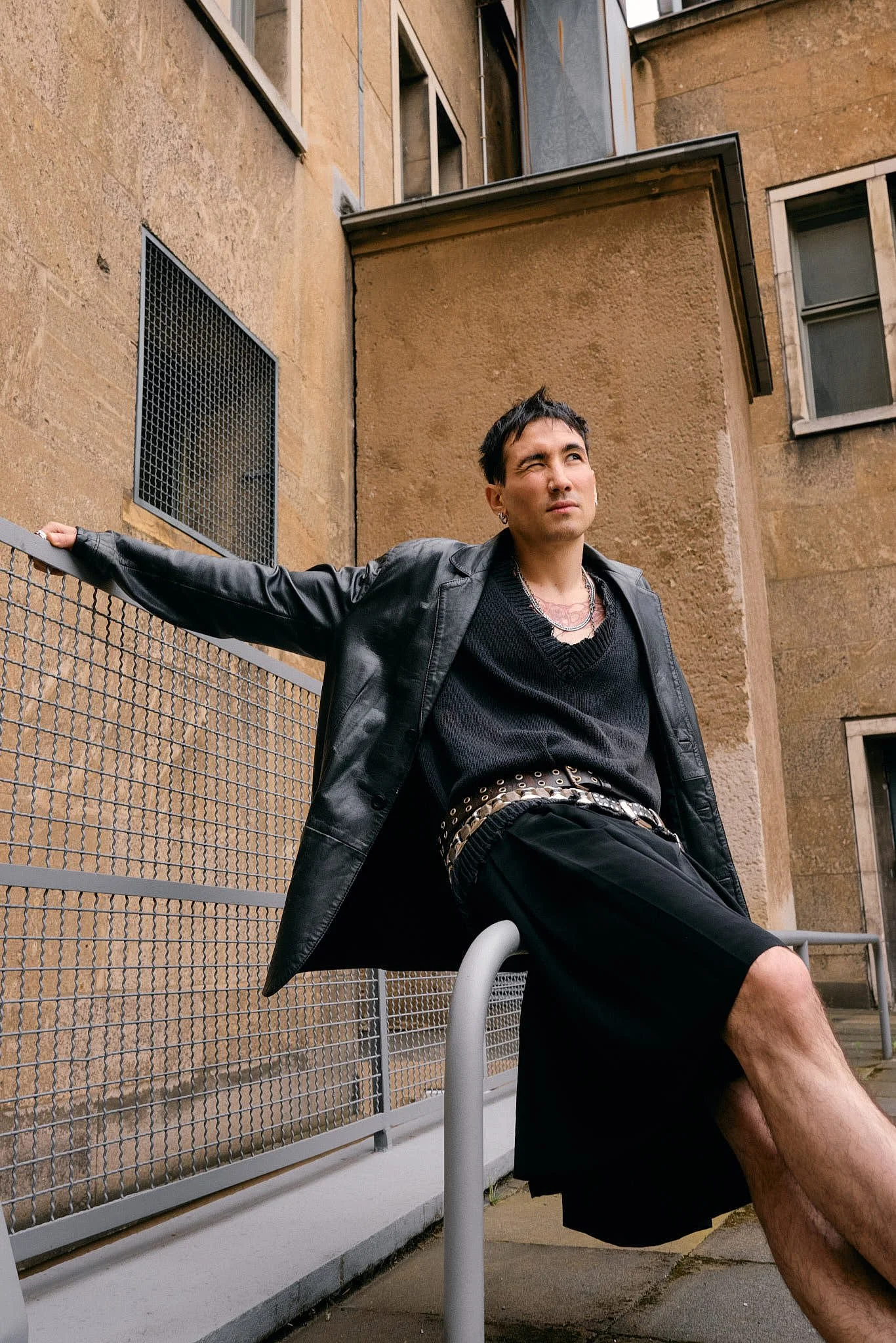 Guest perched on a railing in a black leather blazer, knitted V-neck vest, double eyelet-studded belts and wide black tailored shorts, Berlin Fashion Week SS27 