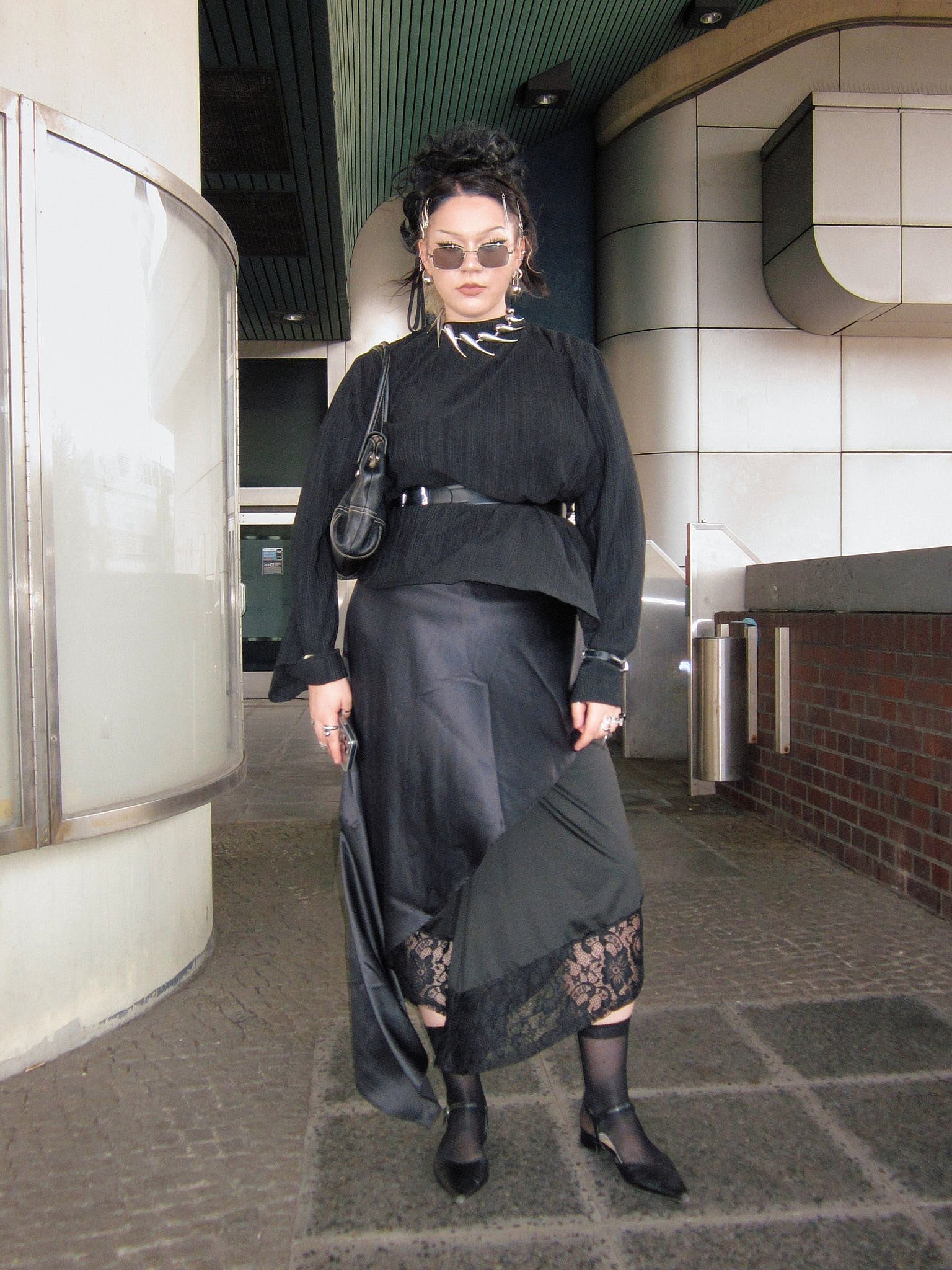 Black crinkled blouse belted over a satin bias skirt with lace hem, spiked silver collar, sheer socks and pointed flats, dark balletcore, outside a station entrance, Berlin Fashion Week SS27 