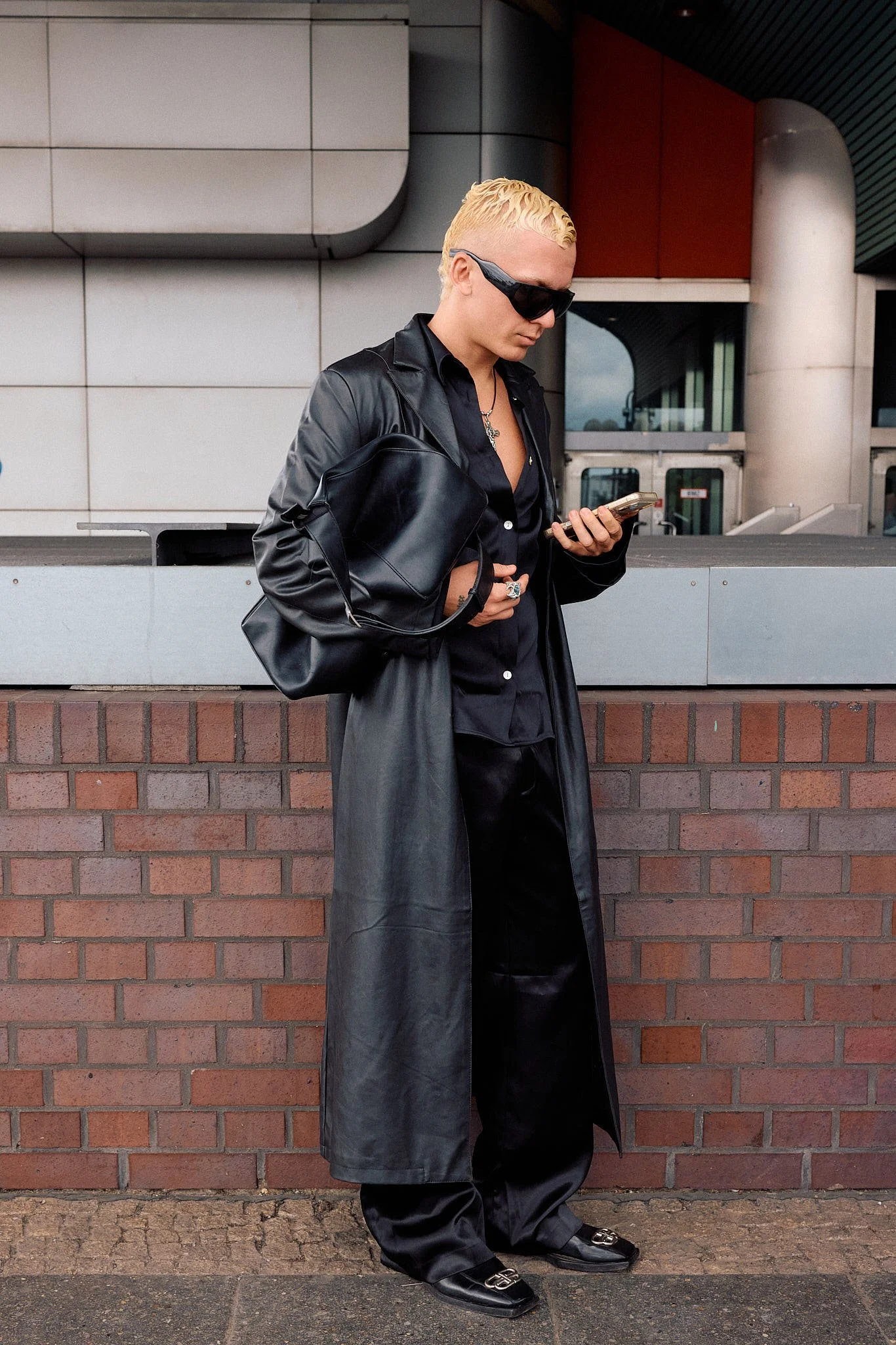  Full-length black leather trench over an open satin shirt and matching trousers, logo-buckle loafers and a slouchy leather shoulder bag, Berlin Fashion Week SS27 