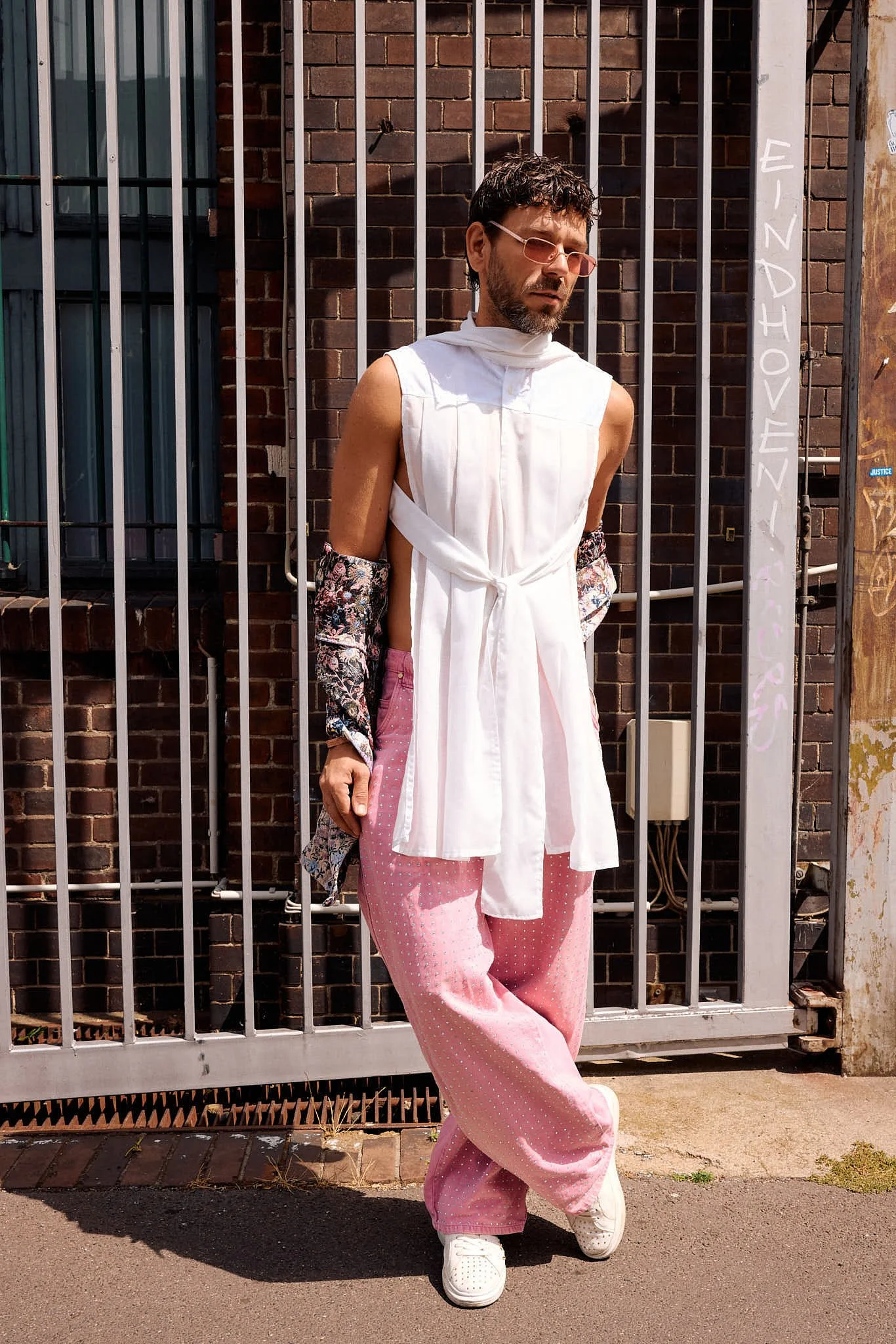  Sleeveless white pleated tunic tied at the waist, worn with floral printed sleeves and pink rhinestone wide-leg jeans against a steel gate, Berlin Fashion Week SS27 