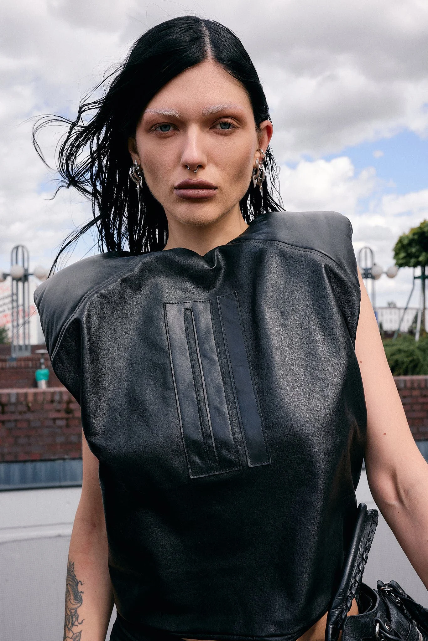  Close-up of a padded-shoulder black leather top with topstitched panel detail, stacked silver ear piercings and a septum ring, shot against Berlin rooftops, Berlin Fashion Week SS27 