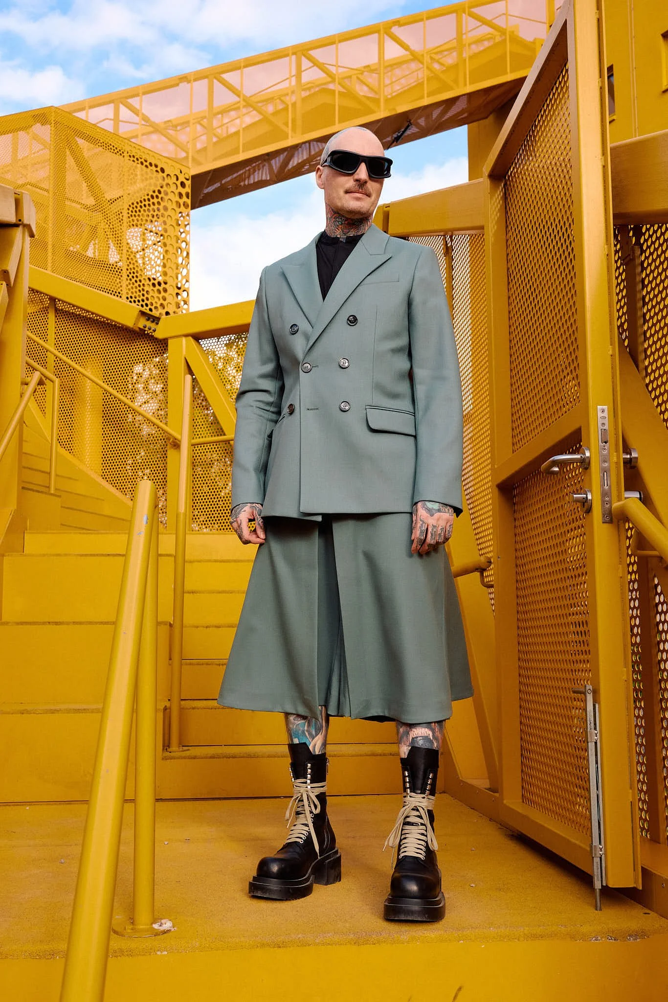  Sage green double-breasted blazer and matching wide pleated culotte shorts with chunky black lace-up platform boots, tailored menswear, on a yellow steel gangway, Berlin Fashion Week SS27 