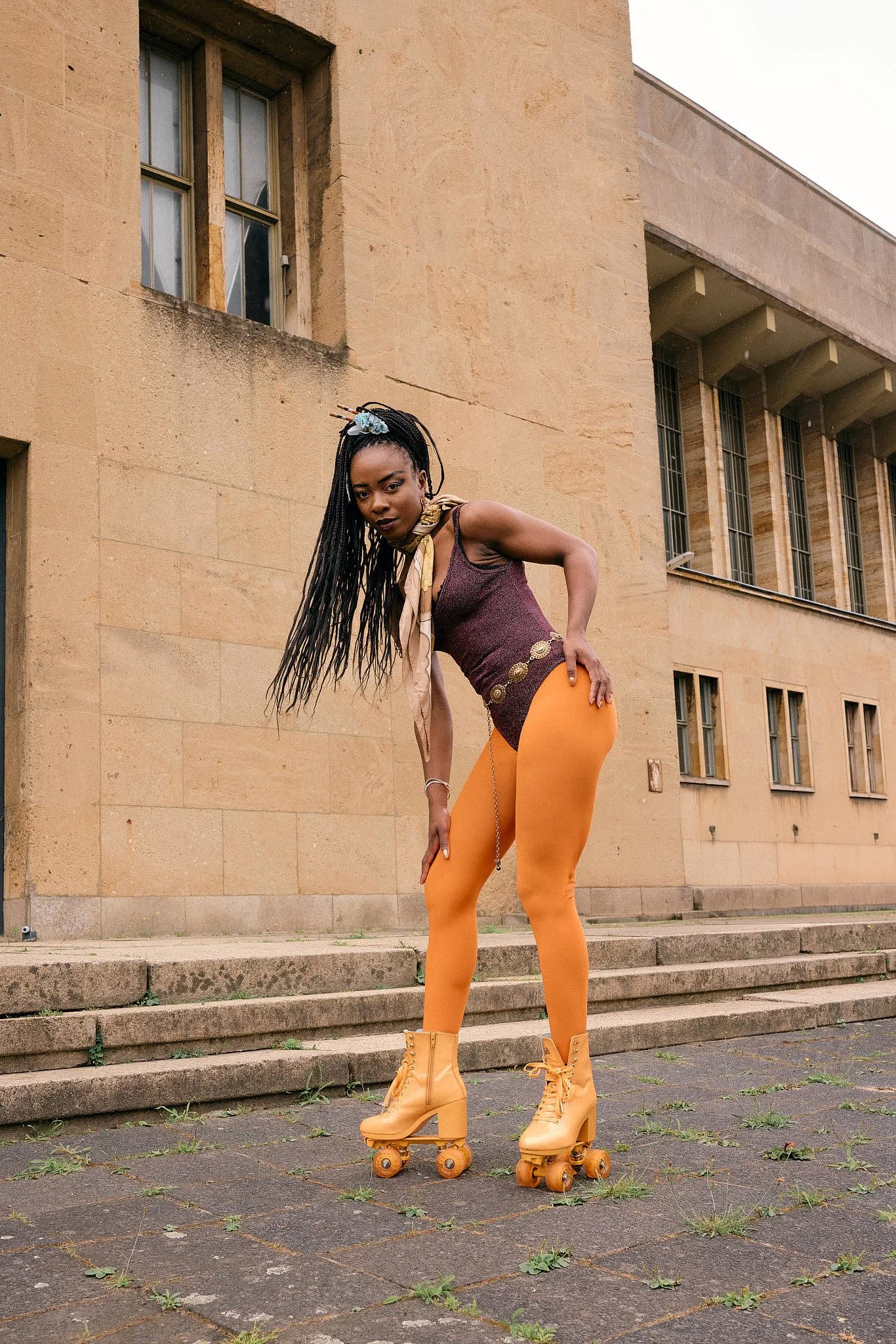  Sport-luxe on wheels: burgundy lurex bodysuit, gold coin belt chain, tangerine tights and matching block-heel roller skates on the limestone steps of a monumental Berlin building, Berlin Fashion Week SS27 