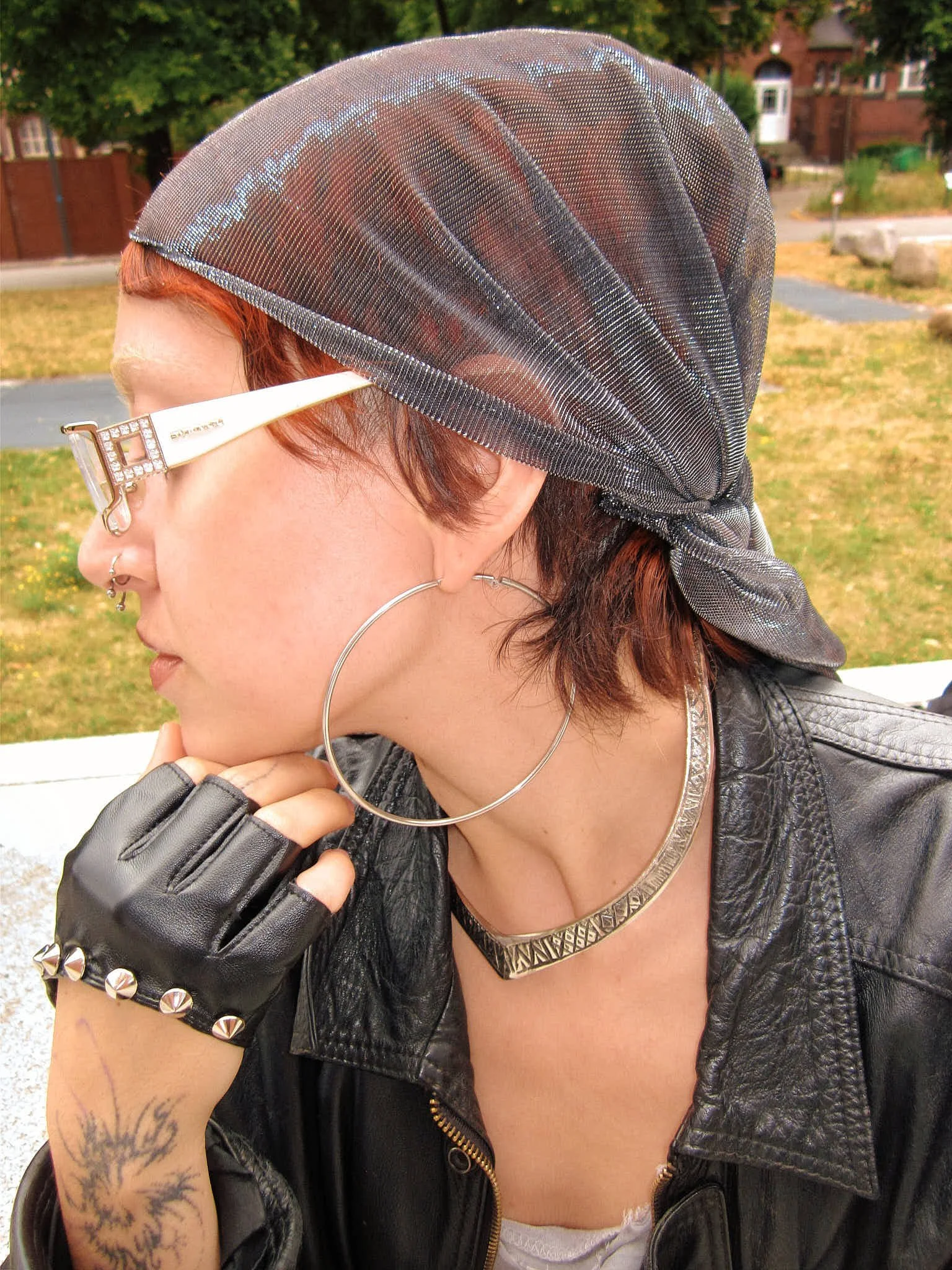  Profile portrait: silver mesh headscarf with rhinestone-trimmed clear glasses, oversized hoops, an engraved silver collar, studded cuff and a black leather jacket, Berlin Fashion Week SS27 