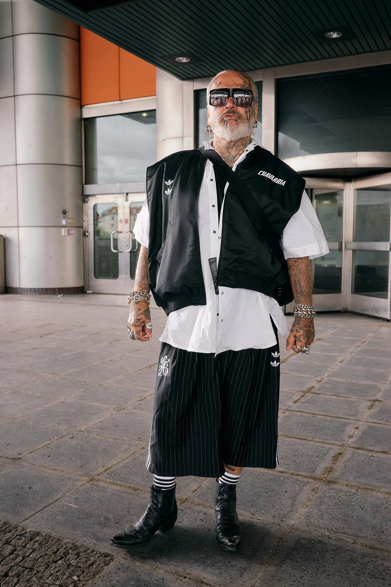  Sport-luxe menswear: sleeveless black adidas bomber printed CHAVARRIA over an oversized white shirt, pinstriped wide adidas shorts, striped socks and black cowboy boots, Berlin Fashion Week SS27 