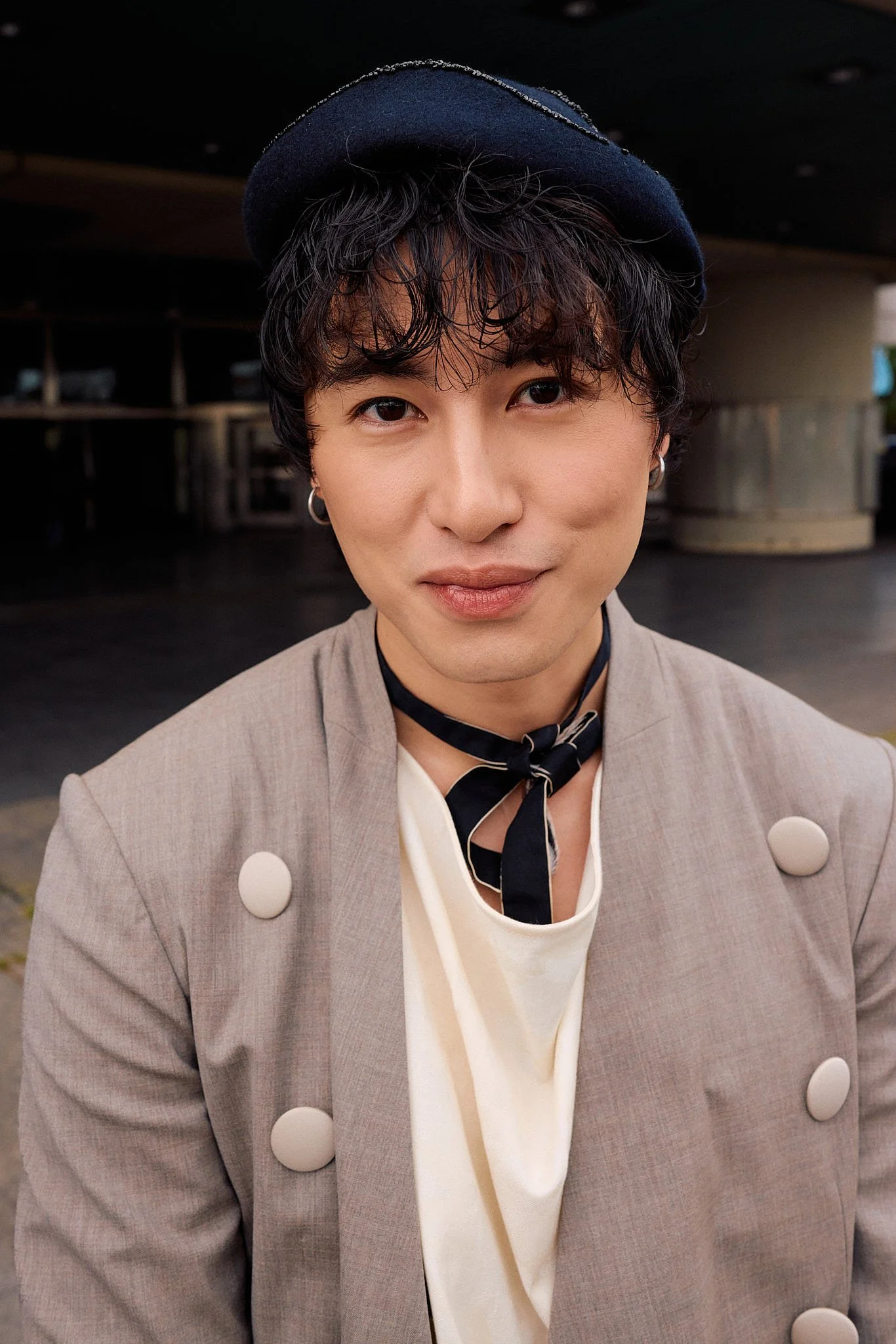  Portrait: navy wool beret and silk neck scarf with a taupe double-breasted blazer with oversized cream buttons over a draped cream top, menswear tailoring, at a show entrance, Berlin Fashion Week SS27 