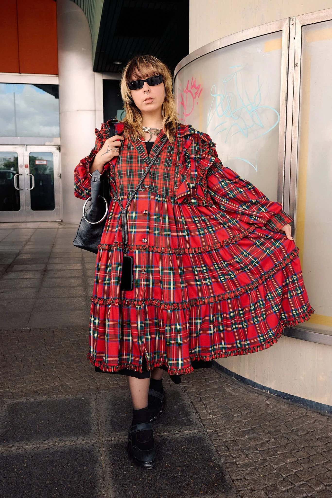  Red tartan tiered ruffle dress with a silver chain choker, black socks and loafers, thrift flip tartan styling, outside a graffiti-marked station entrance, Berlin Fashion Week SS27 