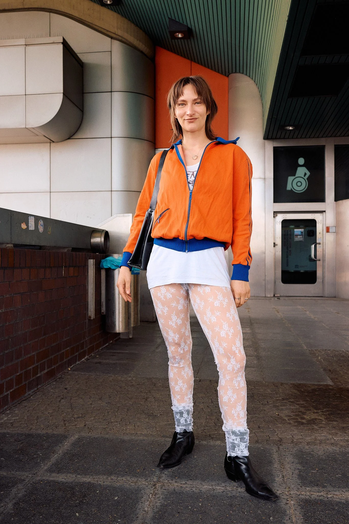  Orange and blue vintage zip-up track jacket over a long white tee with sheer floral lace leggings and black leather ankle boots, retro sportswear layering at a Berlin S-Bahn entrance, Berlin Fashion Week SS27 