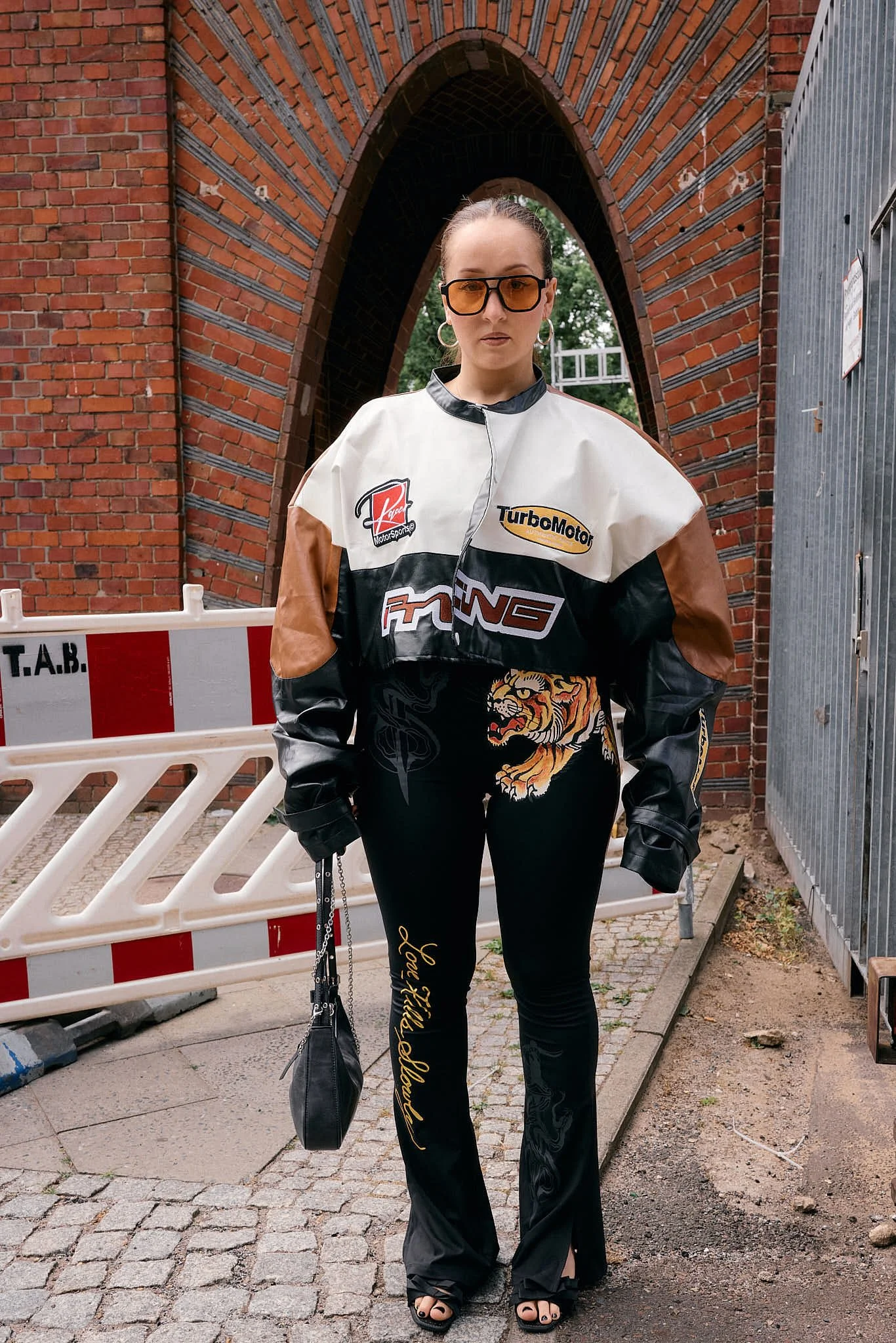  Cropped leather motorsport jacket with TurboMotor racing patches over flared trousers with a tiger graphic and script embroidery, retro sportswear meets thrift flip, beneath a brick arch, Berlin Fashion Week SS27 