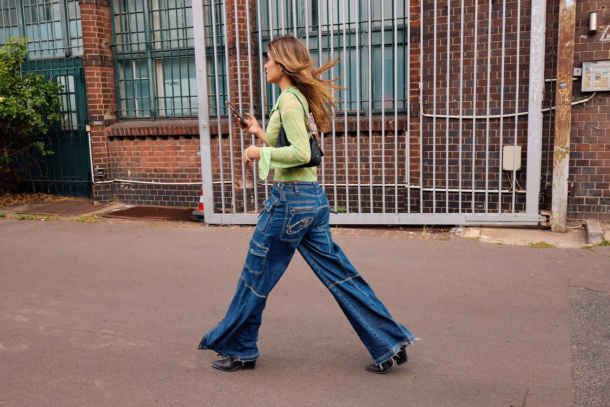  Mid-stride in a sheer lime green top with wide-leg indigo cargo flares, frayed hems and a chain-strap shoulder bag, walking past a brick industrial gate, Berlin Fashion Week SS27 