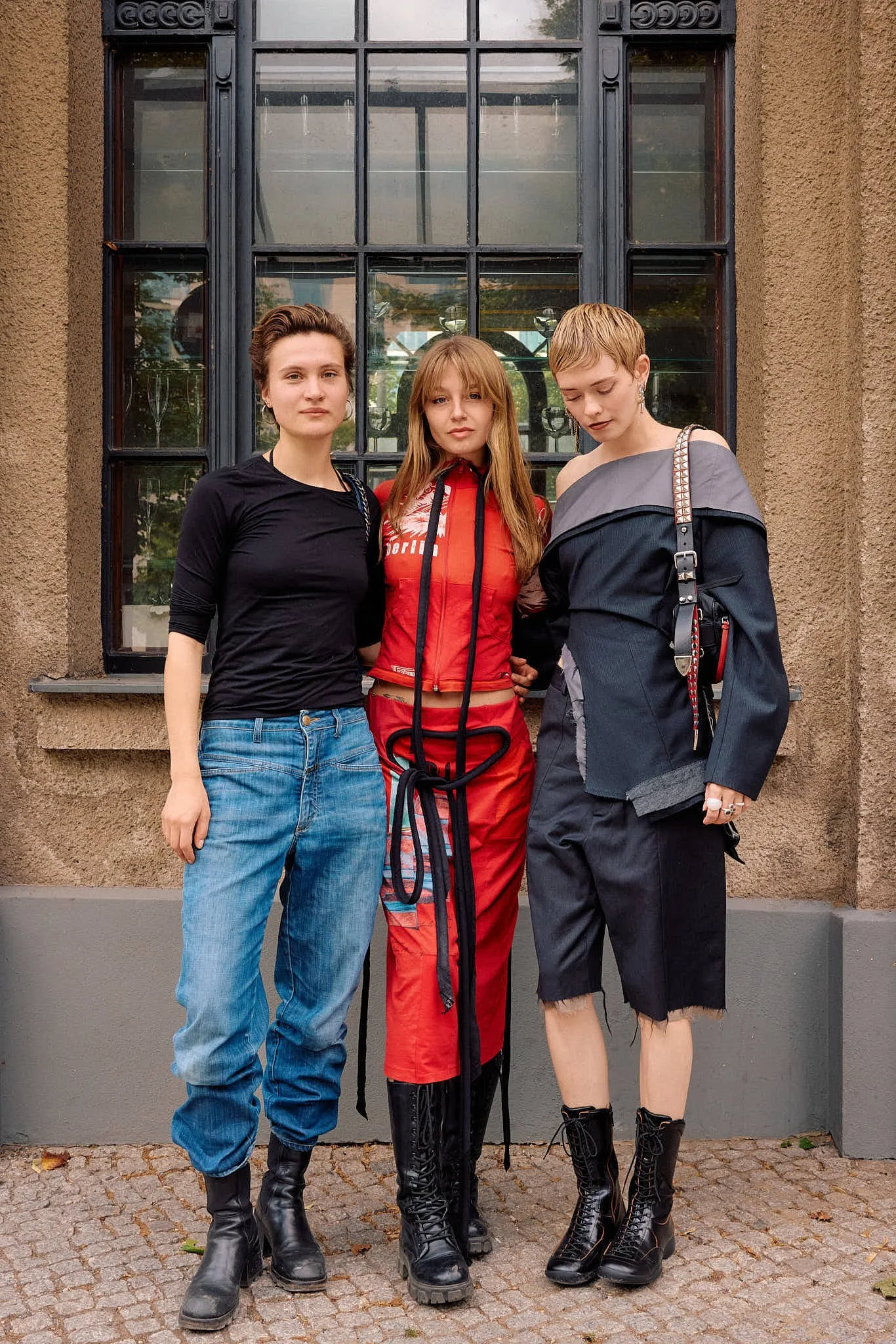  Three guests together: a plain black tee with faded jeans, a red zip-front Berlin print top with matching rope-tied skirt, and a deconstructed off-shoulder tailored jacket with raw-hem shorts, Berlin Fashion Week SS27 