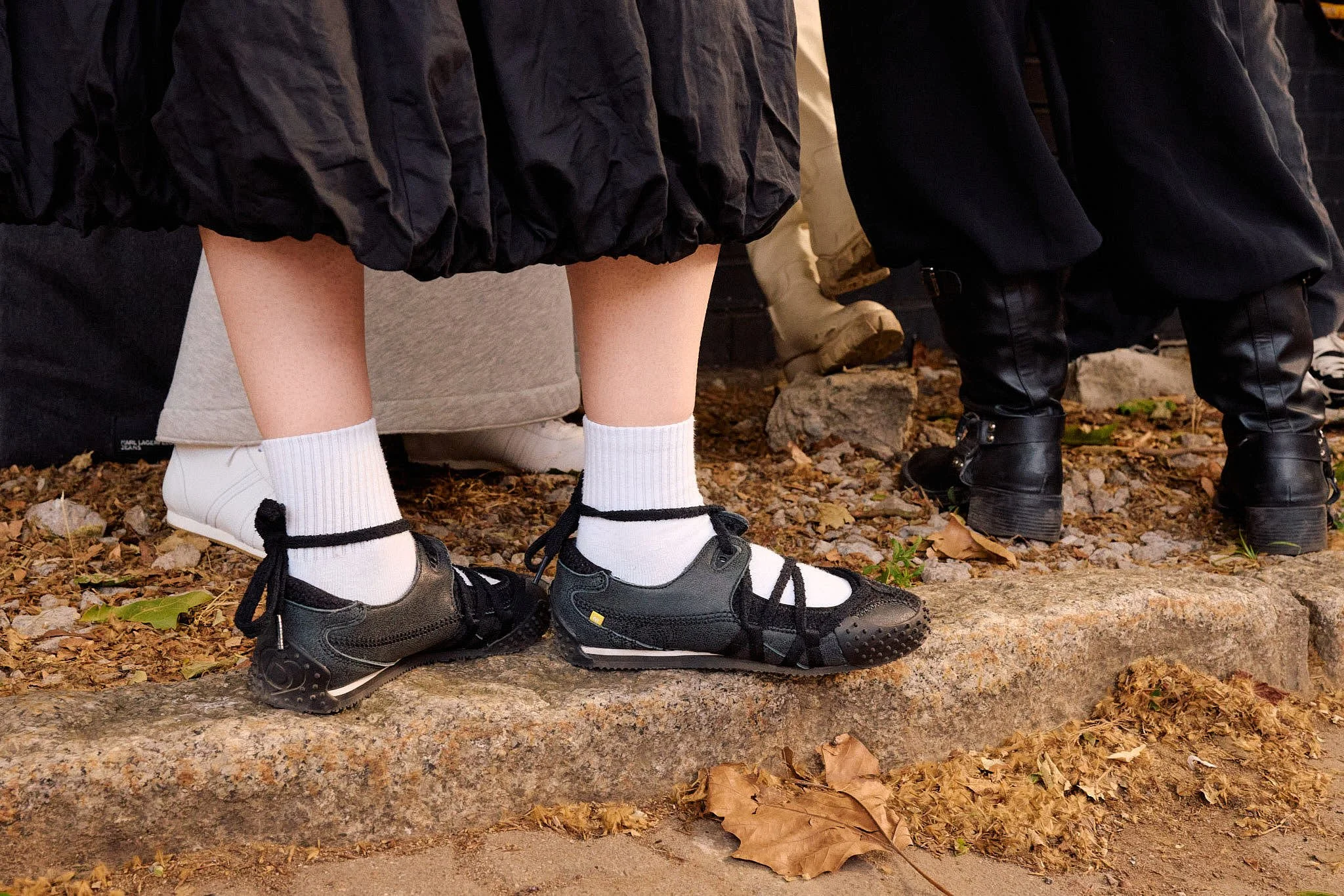  Hybrid footwear detail — black ballet-flat trainers laced with ribbon ties over white sports socks, a dark balletcore favourite spotted kerbside, Berlin Fashion Week SS27 