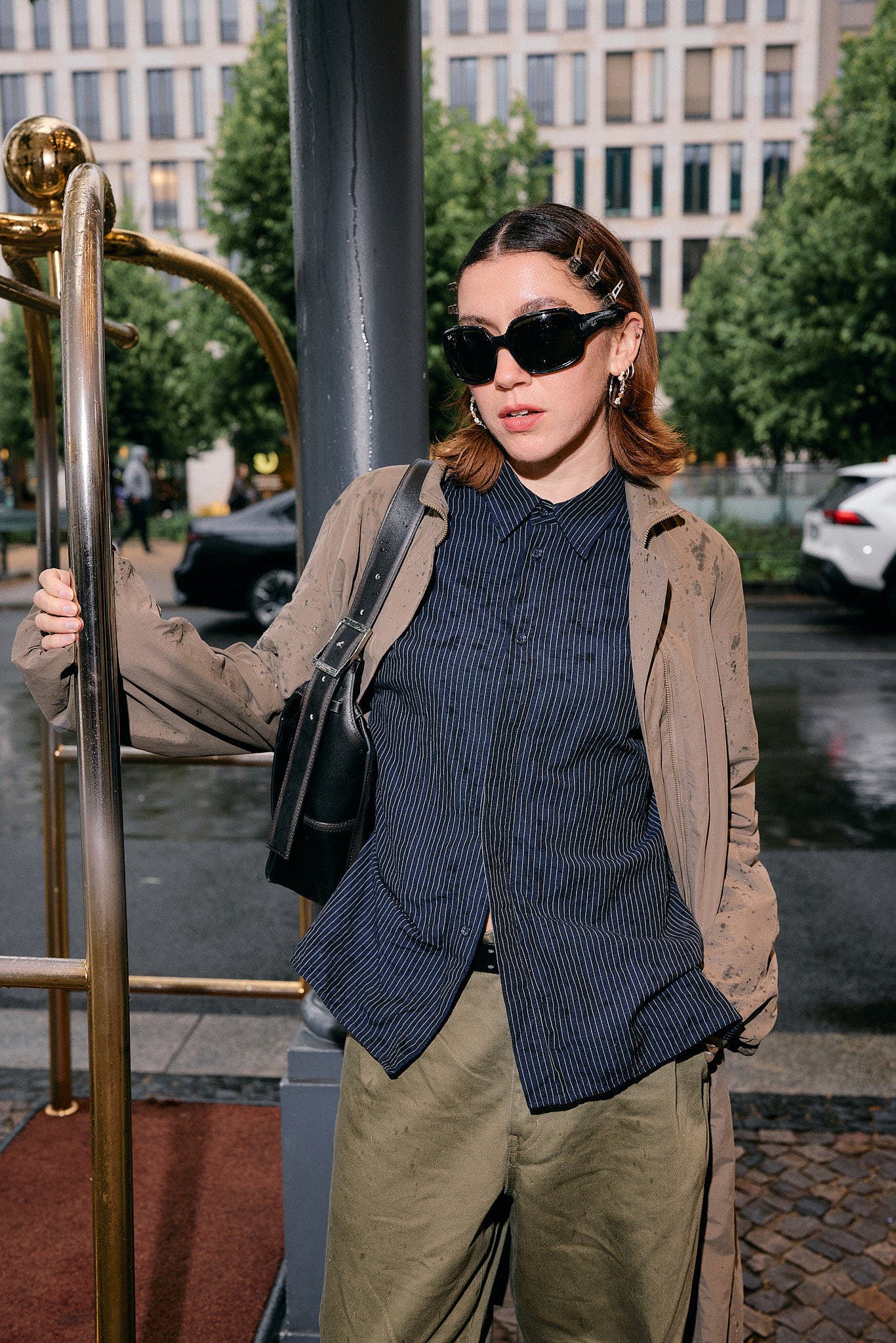  Navy pinstripe shirt under a rain-flecked taupe jacket with olive wide-leg trousers and a black leather shoulder bag, guest on a rainy morning outside a show, Berlin Fashion Week SS27 