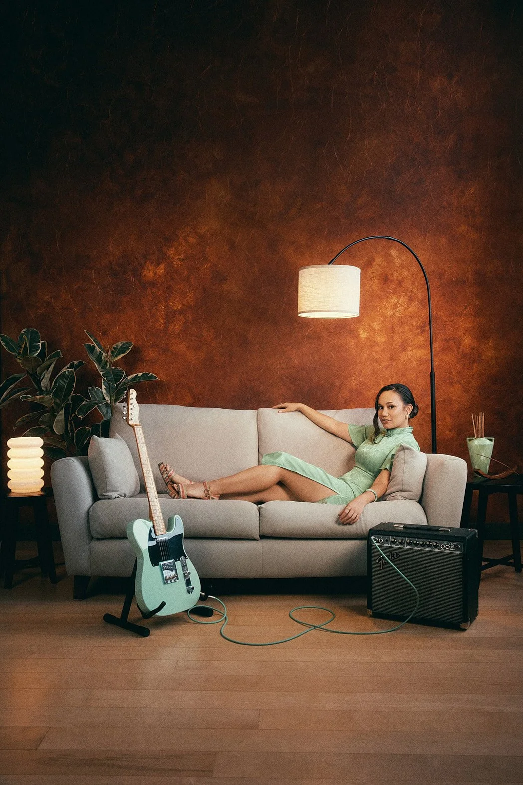  Personal branding photographer London portrait of music artist Denquar laying grey sofa wearing green dress and matching guitar with orange wall and plants. 