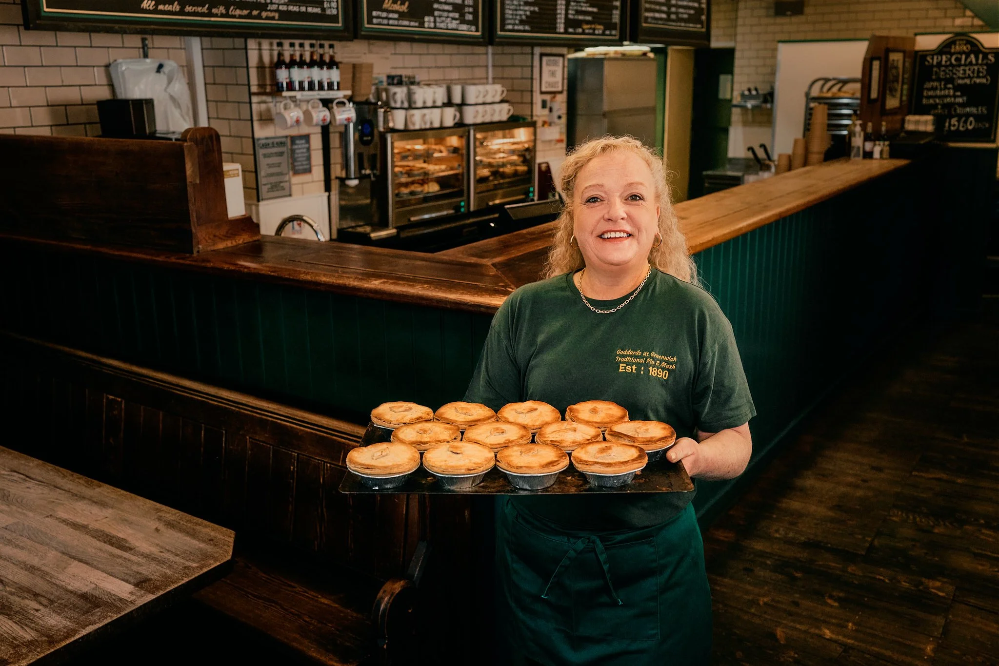 Advertising photography in London for IBM billboard OOH ad campaign for Wimbledon tennis tournament with lifestyle street photography of pie shop worker holding tray of pies in historic shop smiling with countertop and green accents behind her