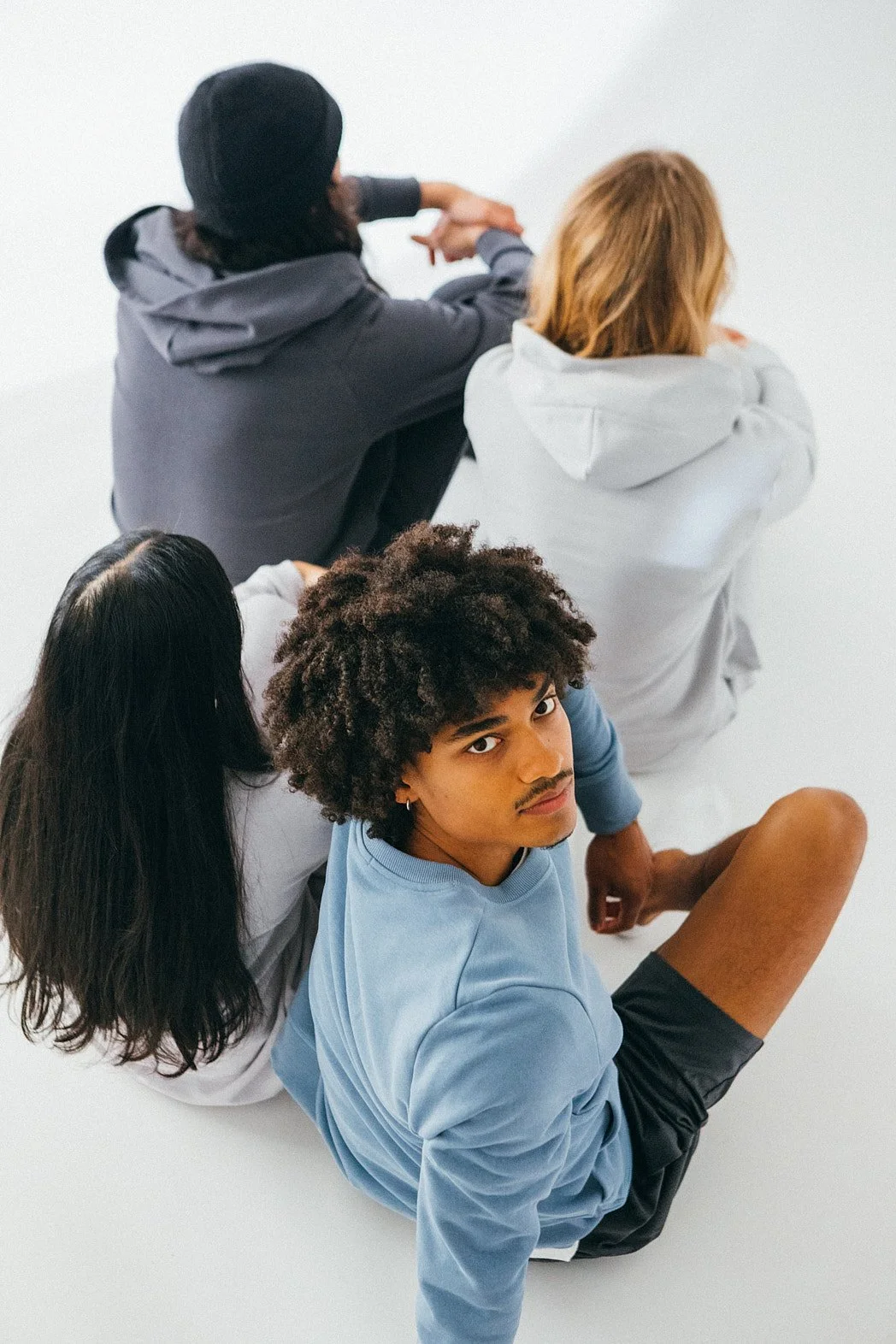 Advertising photographer London Commercial lifestyle image of four models seated on white studio floor with natural light and pastel colour sweatshirt jumpers and one male model with curly hair looks at.