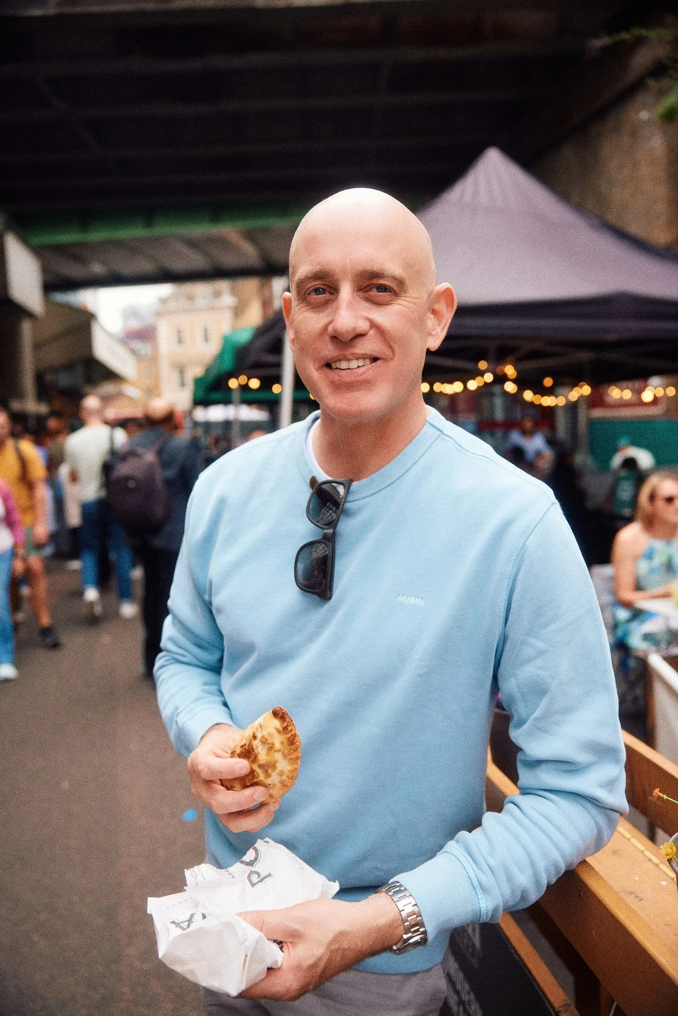 London personal branding photographer Ian Kobylanski captures lifestyle portraits of a bald man enjoying empanada at food market smiling in blue sweater in London