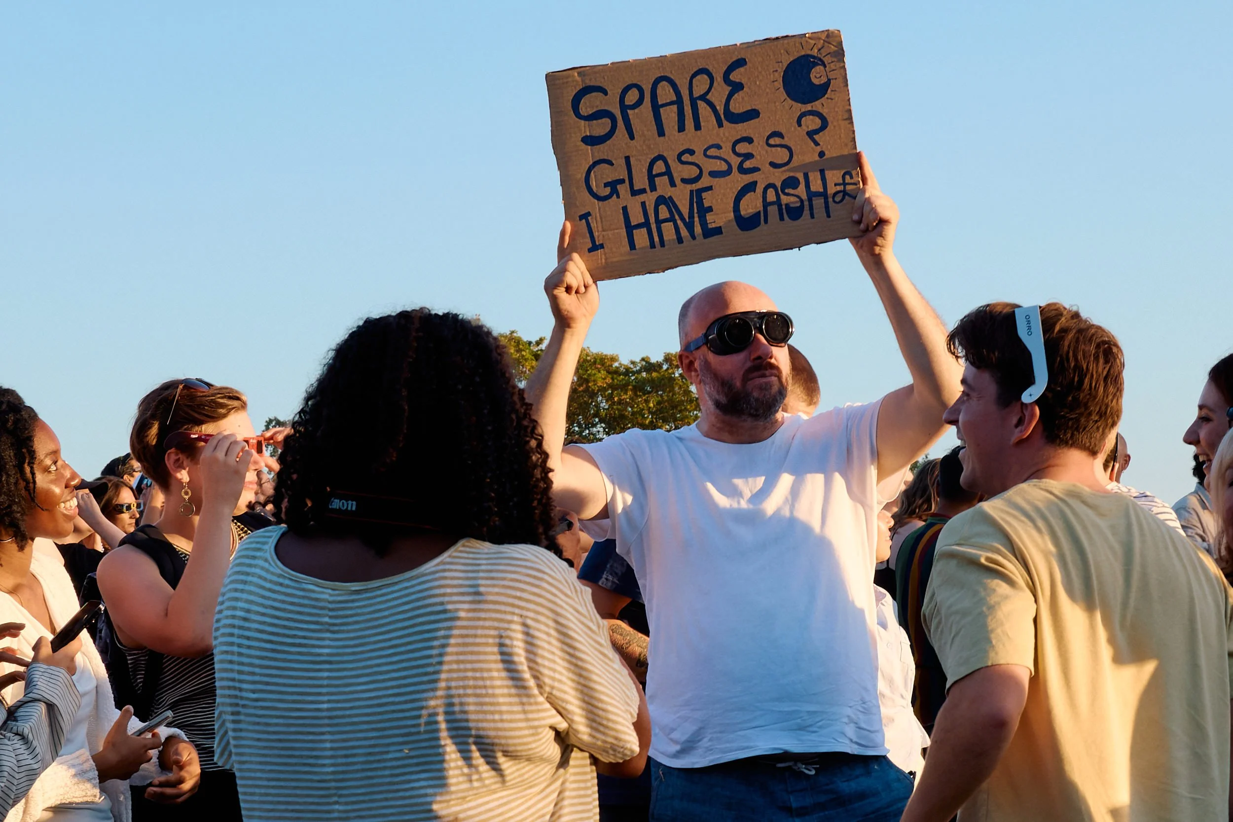 Bald man in sunglasses holding up a cardboard sign reading Spare glasses? I have cash in the crowd on Primrose Hill