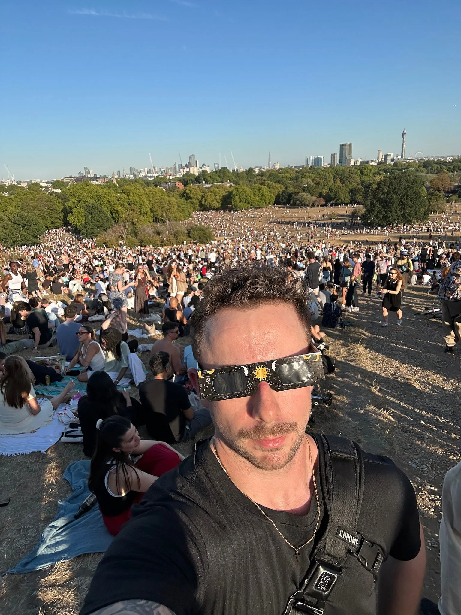 Ian Kobylanski taking a selfie in eclipse glasses at the top of Primrose Hill, the crowd and BT Tower behind him