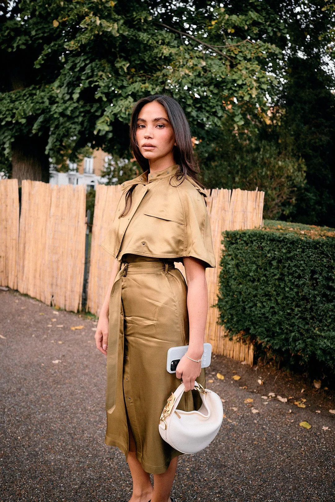 Street style portrait at London Fashion Week of model looking at camera with burberry tan dress, relaxed expression with tree and house background