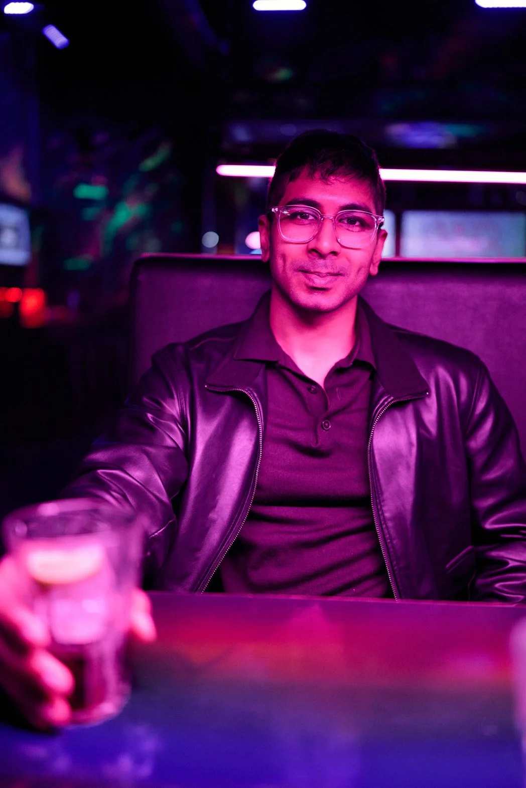 Dating photography lifestyle portrait of man at arcade bar confident smile leather jacket and glasses lit by purple light dark room relaxed