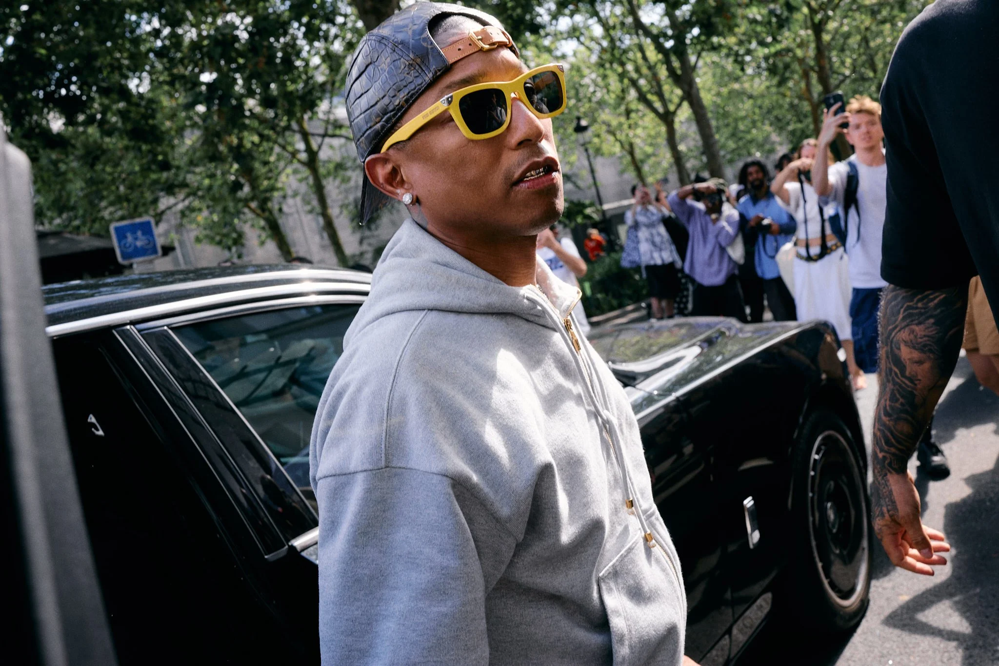 Pharrell Williams in a grey hooded jacket, yellow sunglasses and a Louis Vuitton monogram cap outside the Comme des Garçons show, Paris Fashion Week SS27 menswear