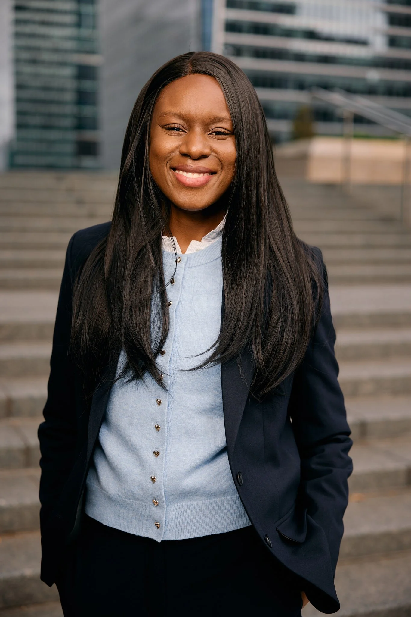 London headshot photographer captures corporate headshot in Canary Wharf of dark skin woman in blue suit smiling at camera with glass building background