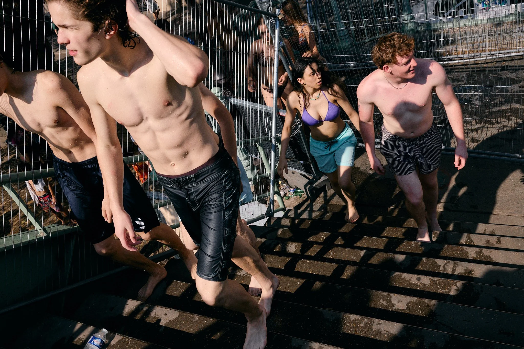 Shirtless young swimmers and a woman in a purple bikini top climb the steps through temporary fencing at Canal Saint-Martin during the Paris heatwave, Paris Fashion Week SS27 menswear