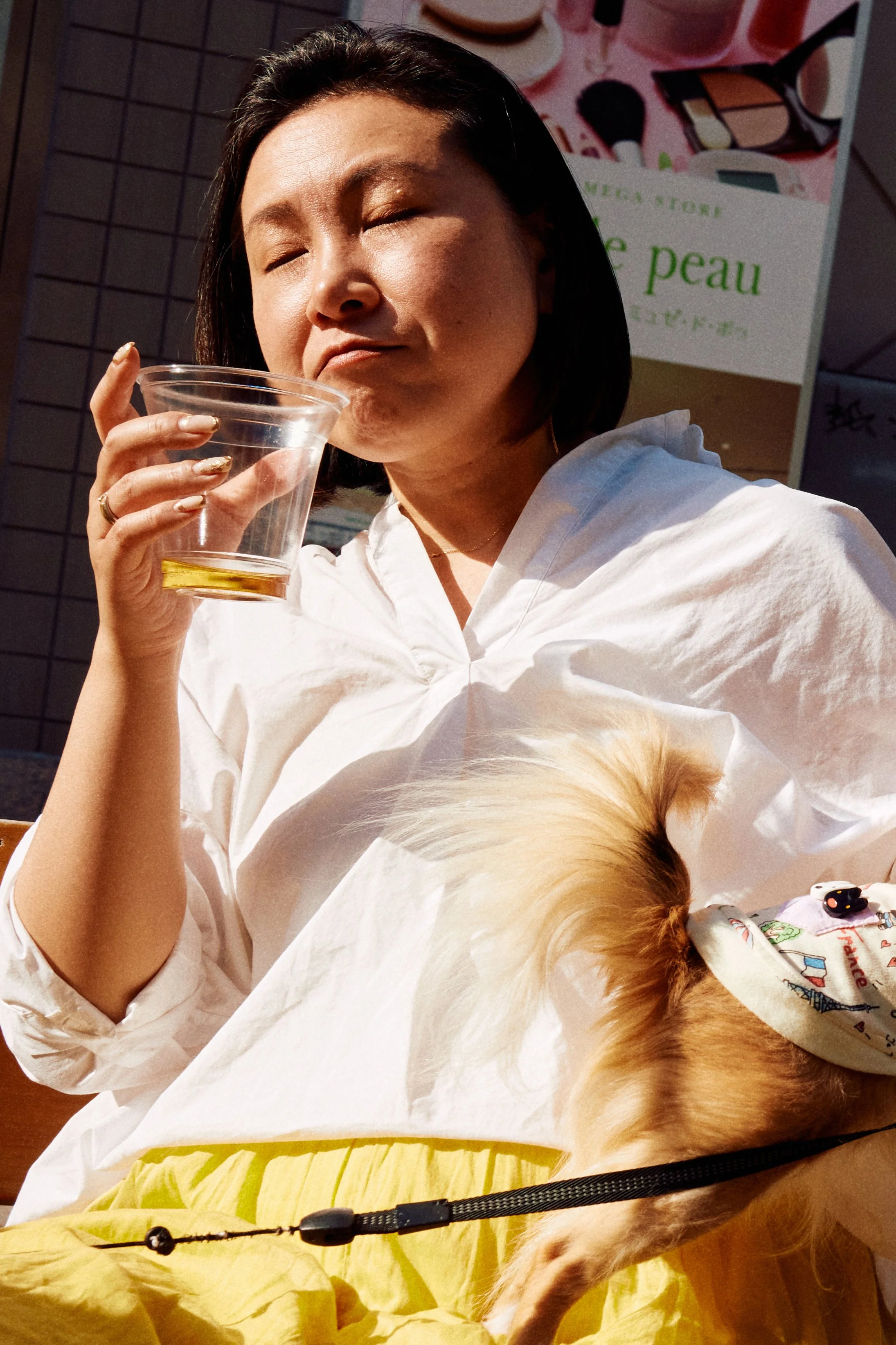 Street photography in Tokyo of woman sipping drink with eyes closed in sun with dog on lap