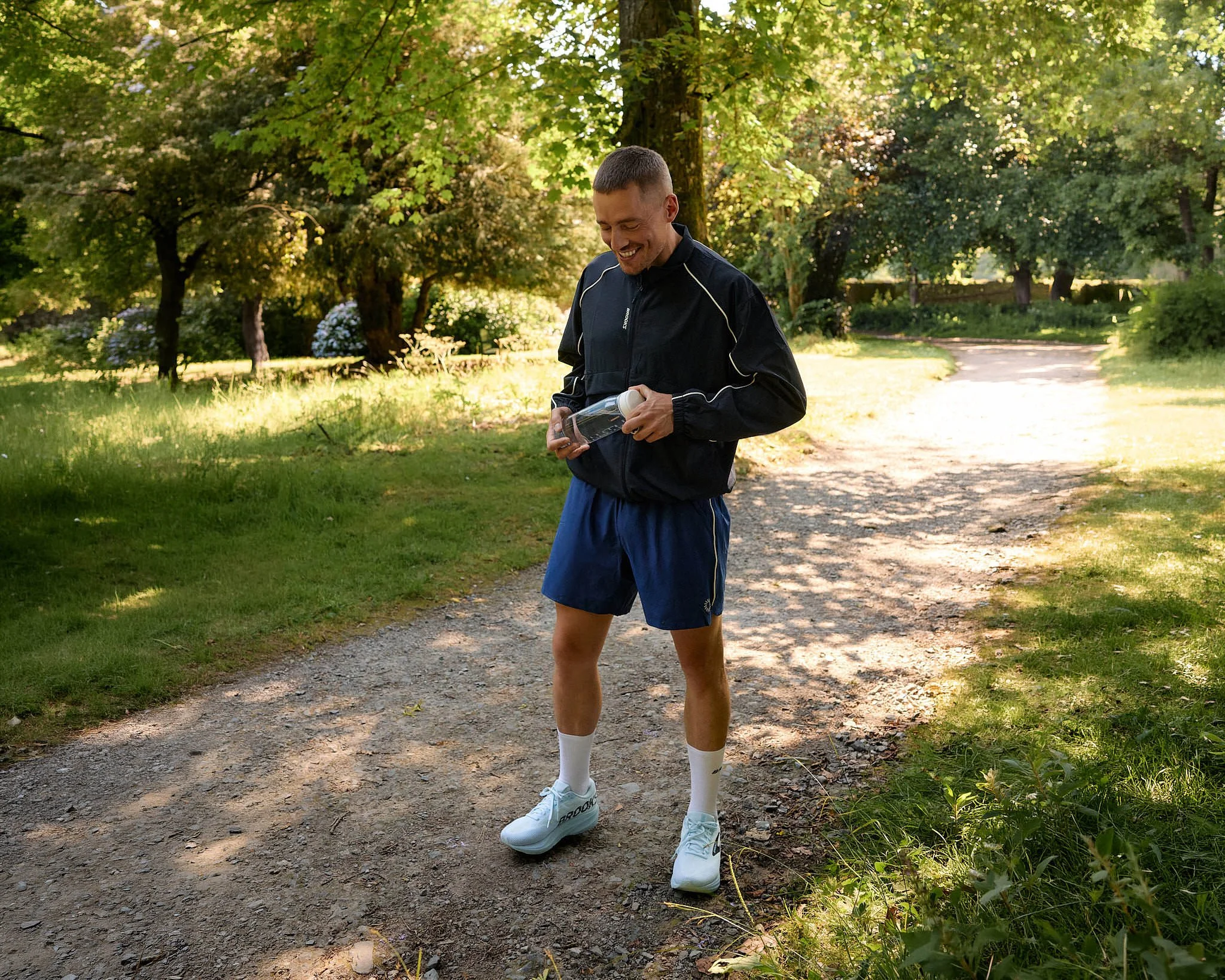 Dermot Kennedy smiling as he opens a water bottle on a shaded woodland path in pale blue Brooks running shoes