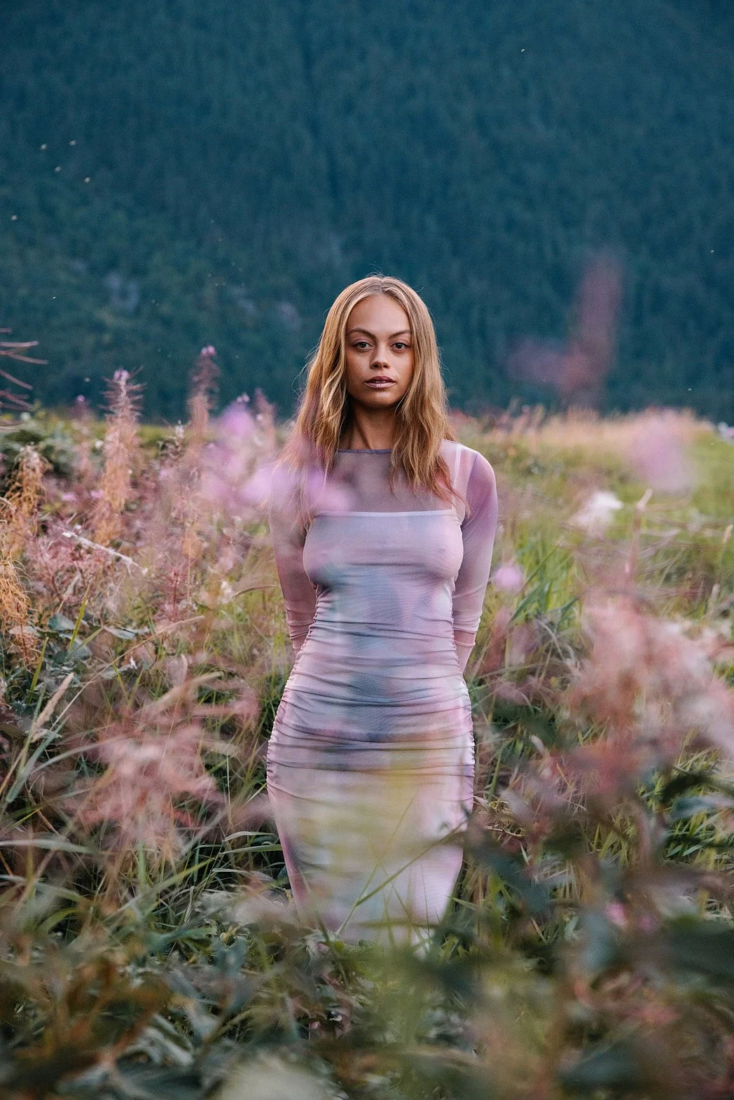 Fashion photographer London editorial image featuring a female model outdoors with forest and mountain background with violet purple dress framed by matching violet flowers and flies in air looking at camera.