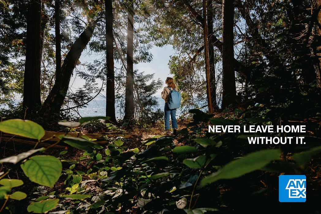 Advertising photographer London campaign image for speculative American Express campaign image of blonde actress lit by sun ray with blue backpack, ocean through trees, and green leaves in foreground.