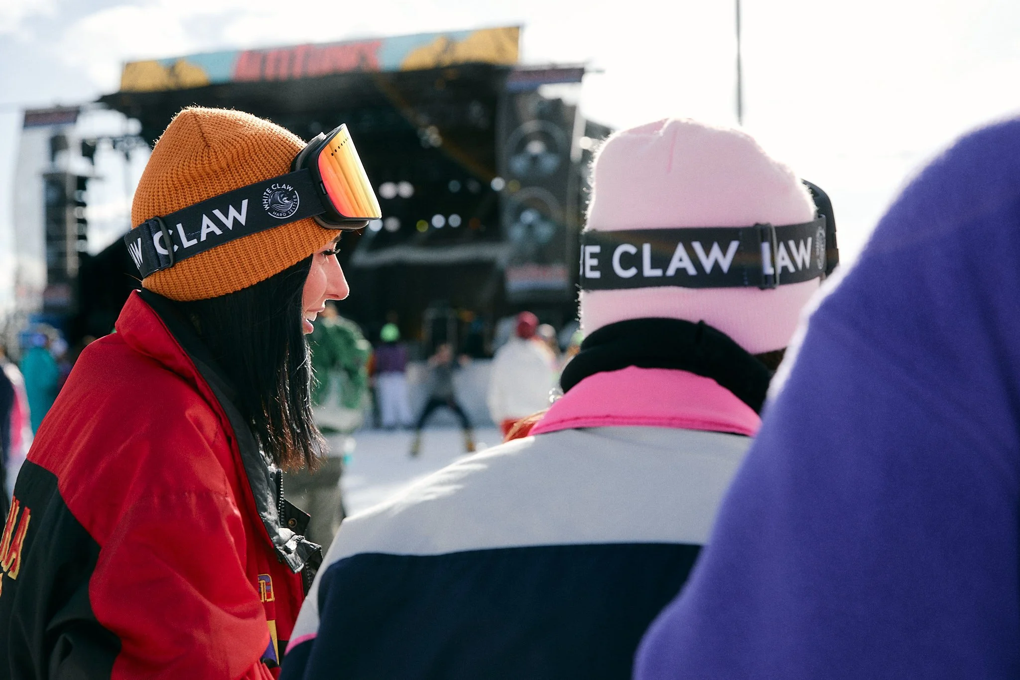 Advertising photographer London brand campaign image of White Claw Hard Seltzer of two women with colourful toques and jackets wearing branded ski goggles smiling and looking off with Altitunes Music.