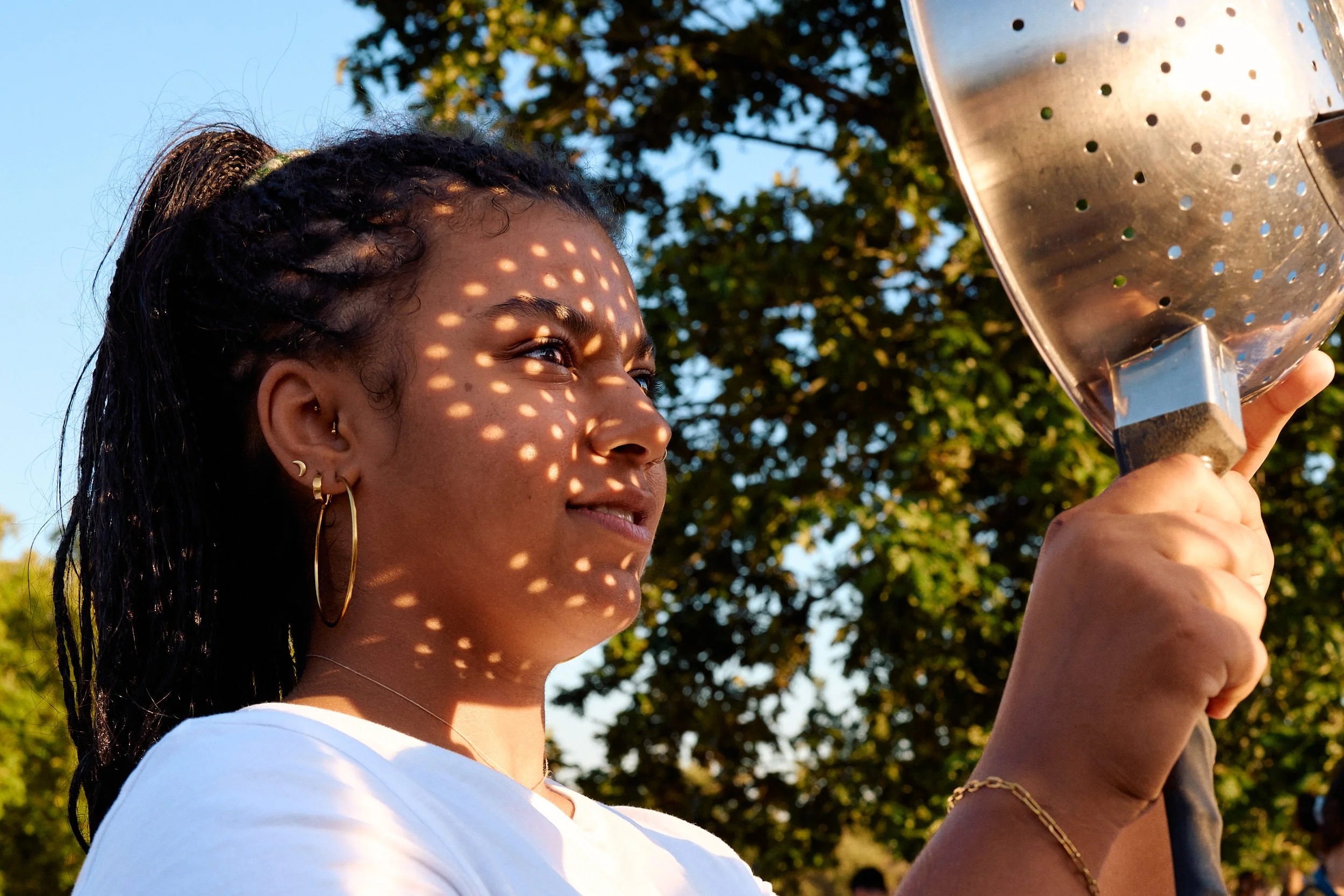 Young woman holding a metal colander up to the sun, dozens of tiny crescent suns projected across her face
