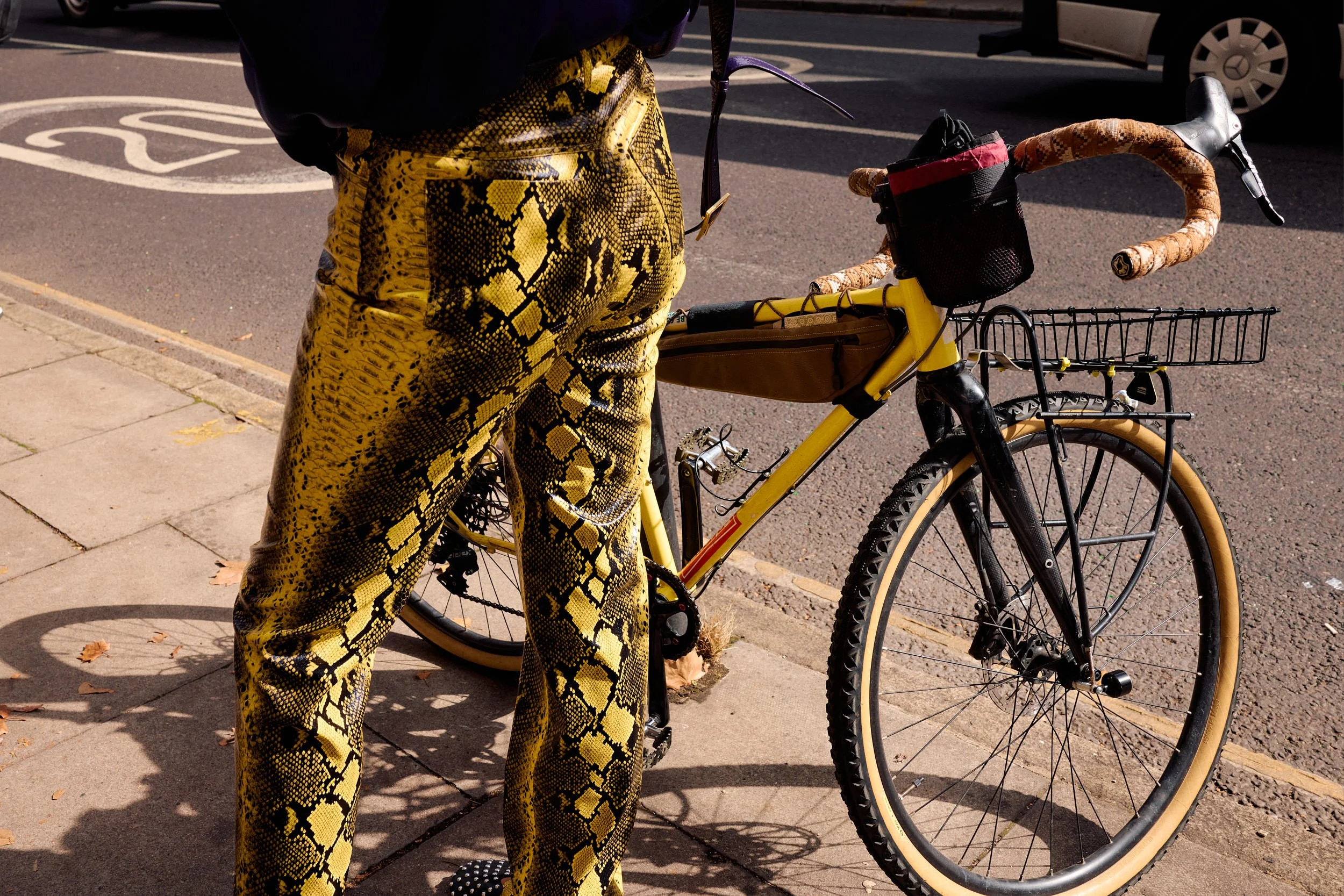 Detail of gold snakeskin-print trousers beside a yellow road bike with a wicker basket on a London street during LFW