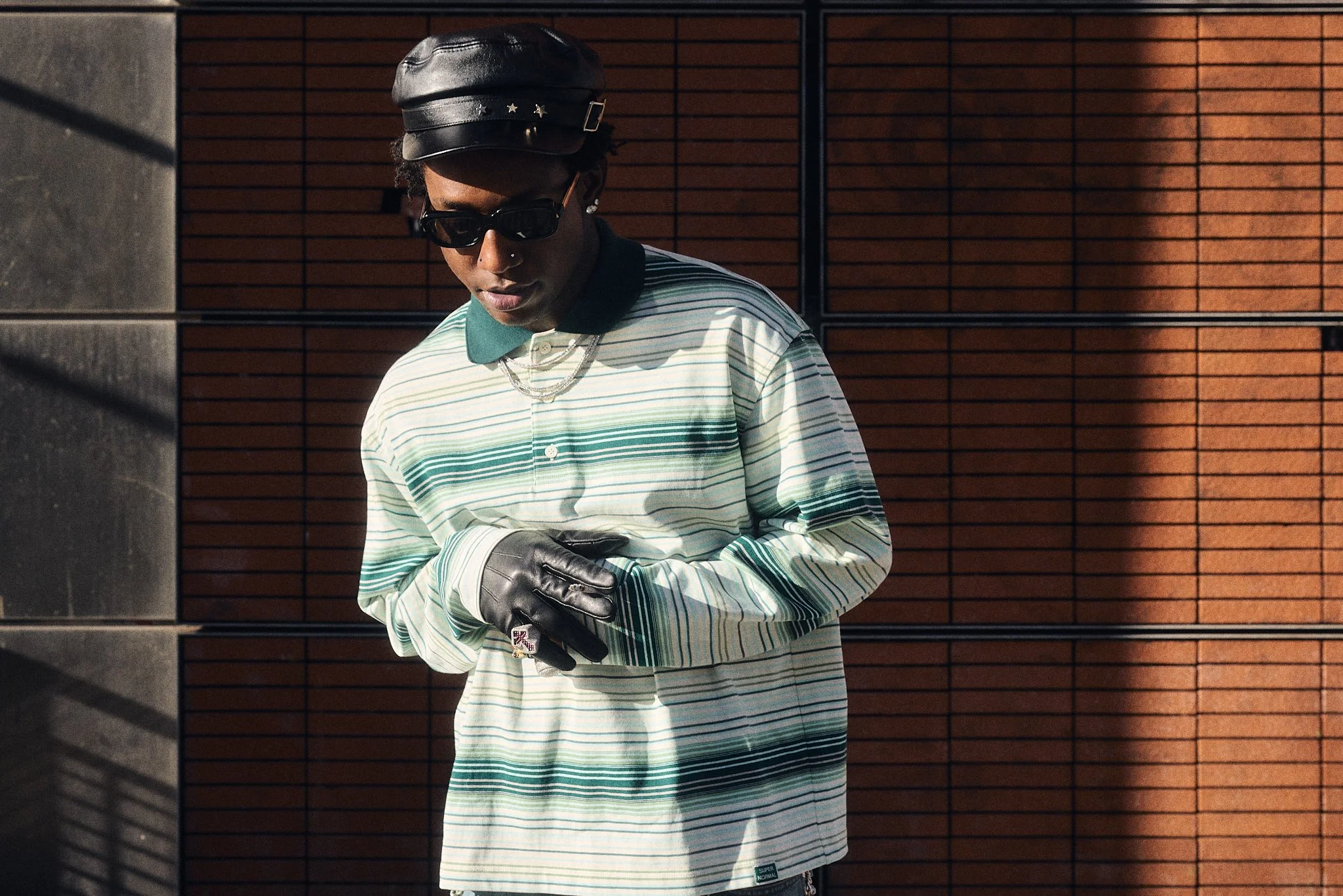 Editoirial fashion photography with Lagos, Nigeria music artist Shallipopi at Paris Fashion Week AW26 street style photoshoot wearing leather hat, sunglasses, green striped rugby shirt, black leather gloves and diamond rings