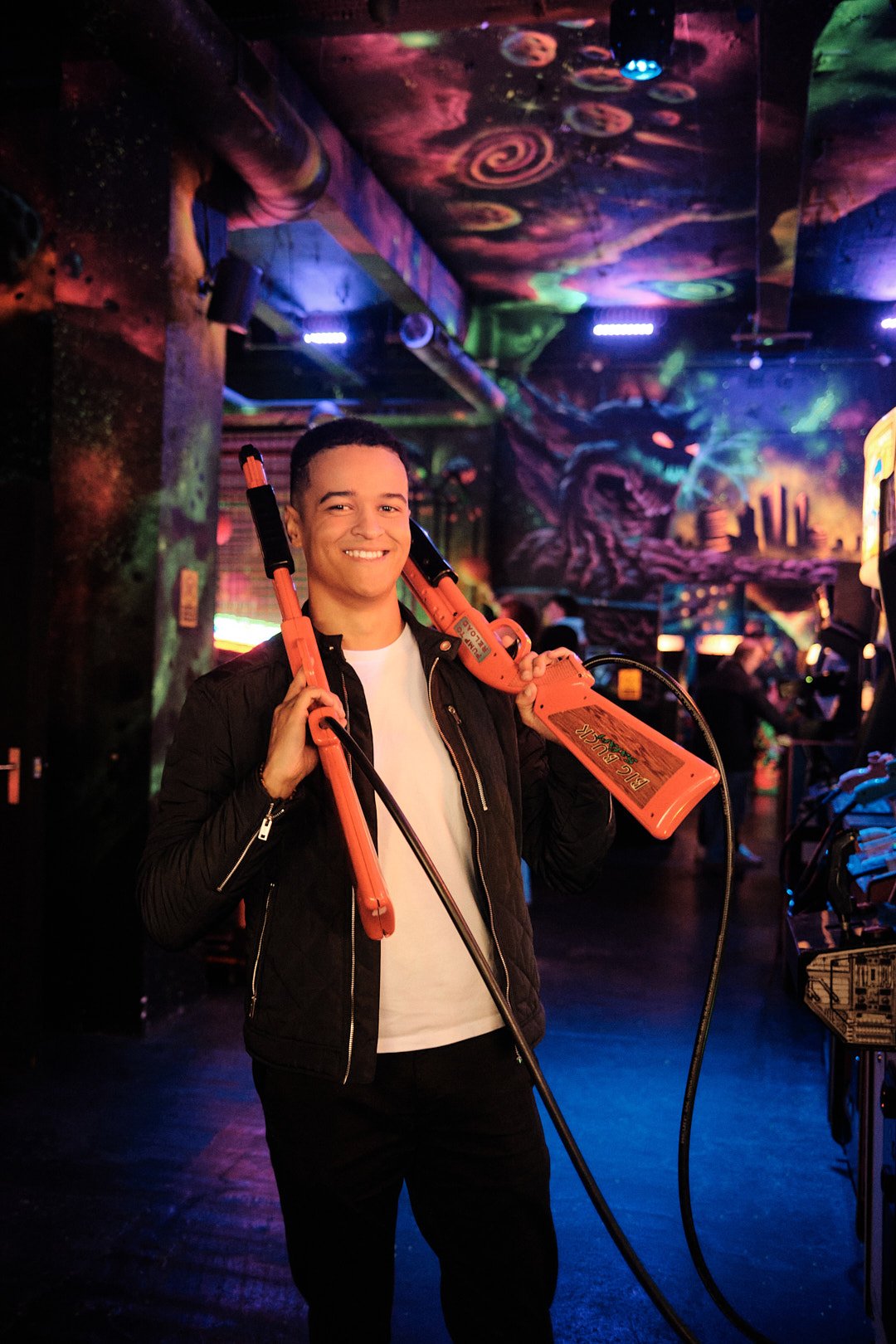 Dating photography in London of light skin man in an arcade holding game shotguns on his shoulder playful portrait