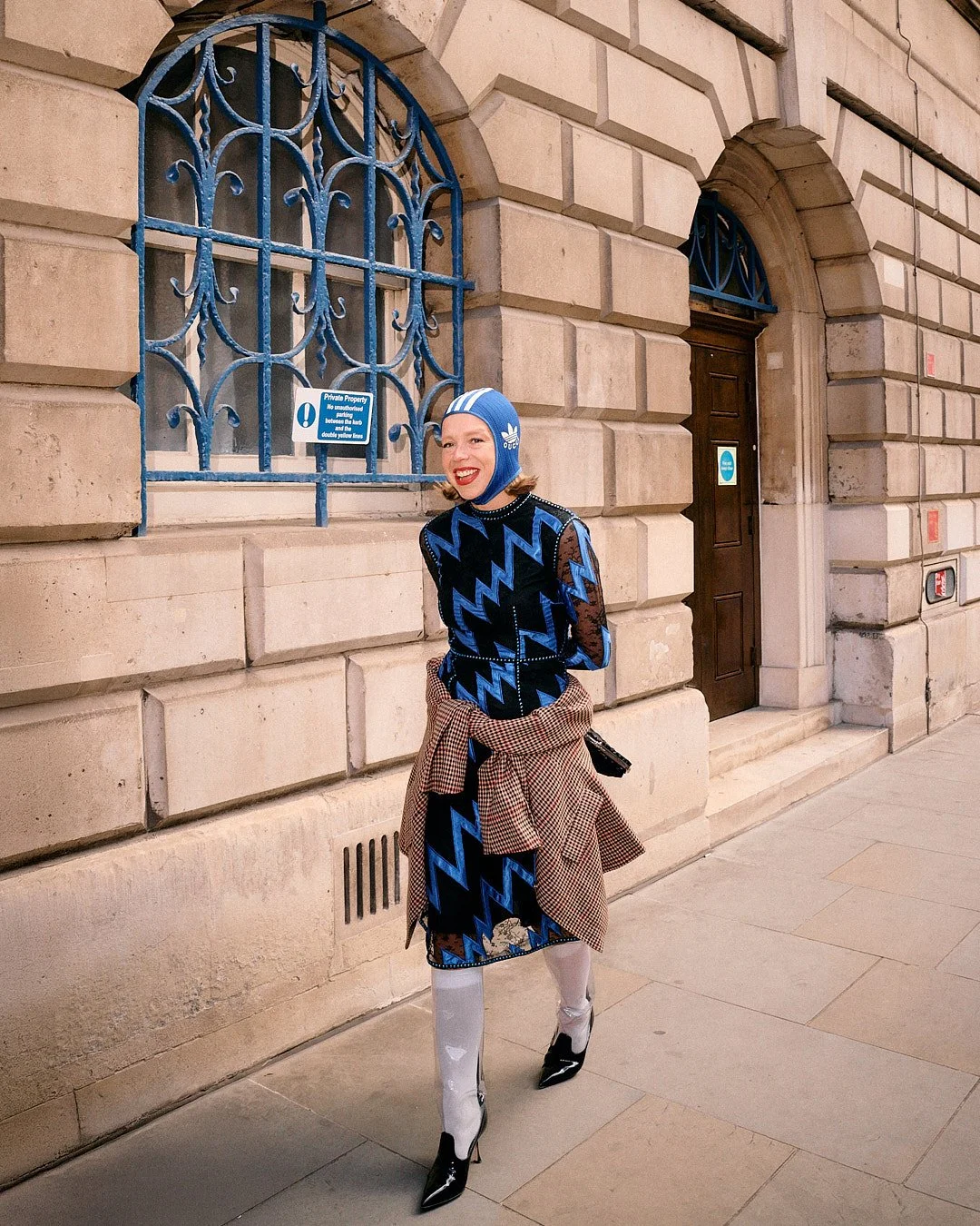 Street style portrait at London Fashion Week of woman wearing blue Gucci Adidas head covering and blue zig zag top in front of historical building