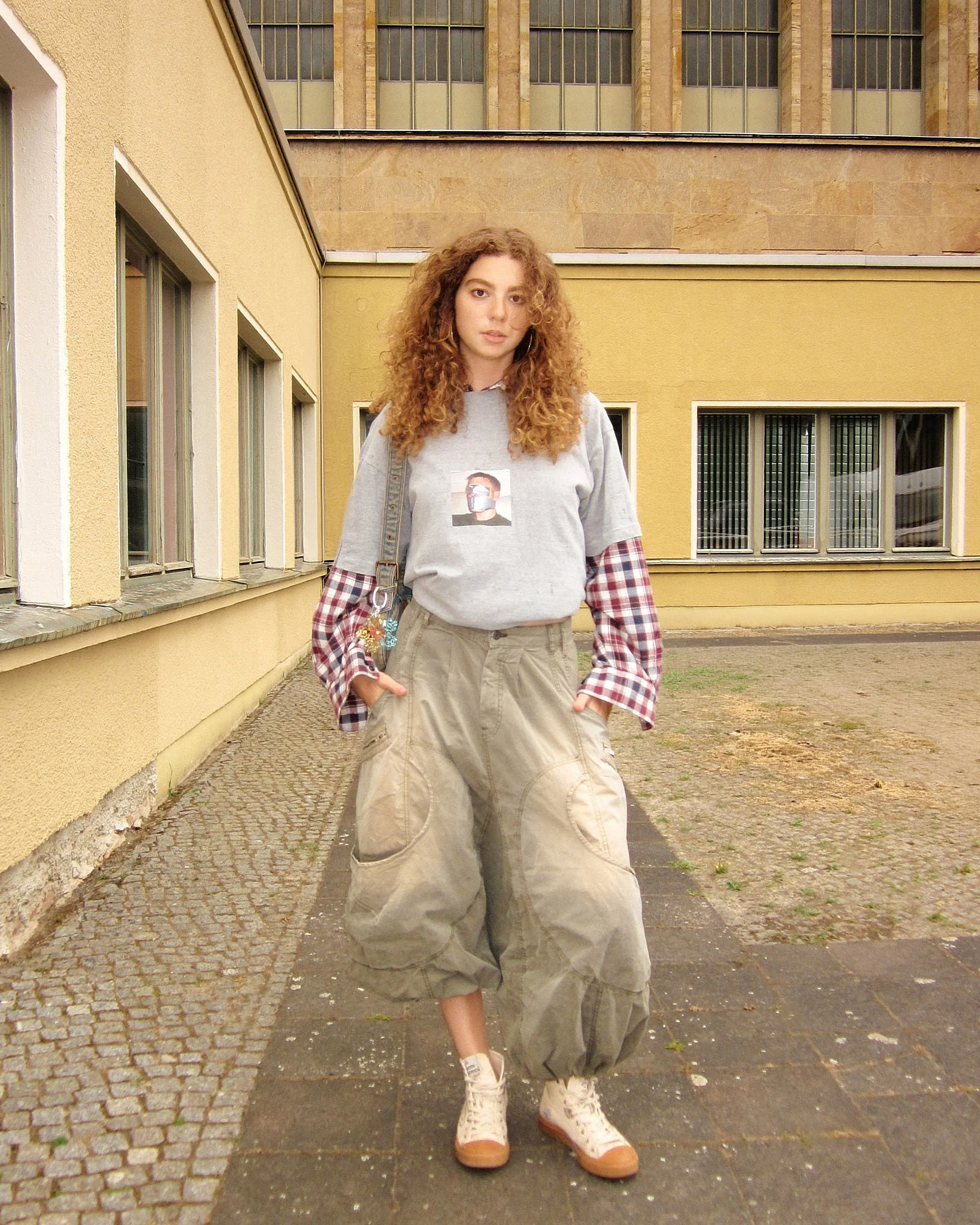 Thrift flip layering — grey photo-print sweatshirt pulled over a red check shirt with oversized khaki parachute cargo trousers and cream trainers, Berlin Fashion Week SS27