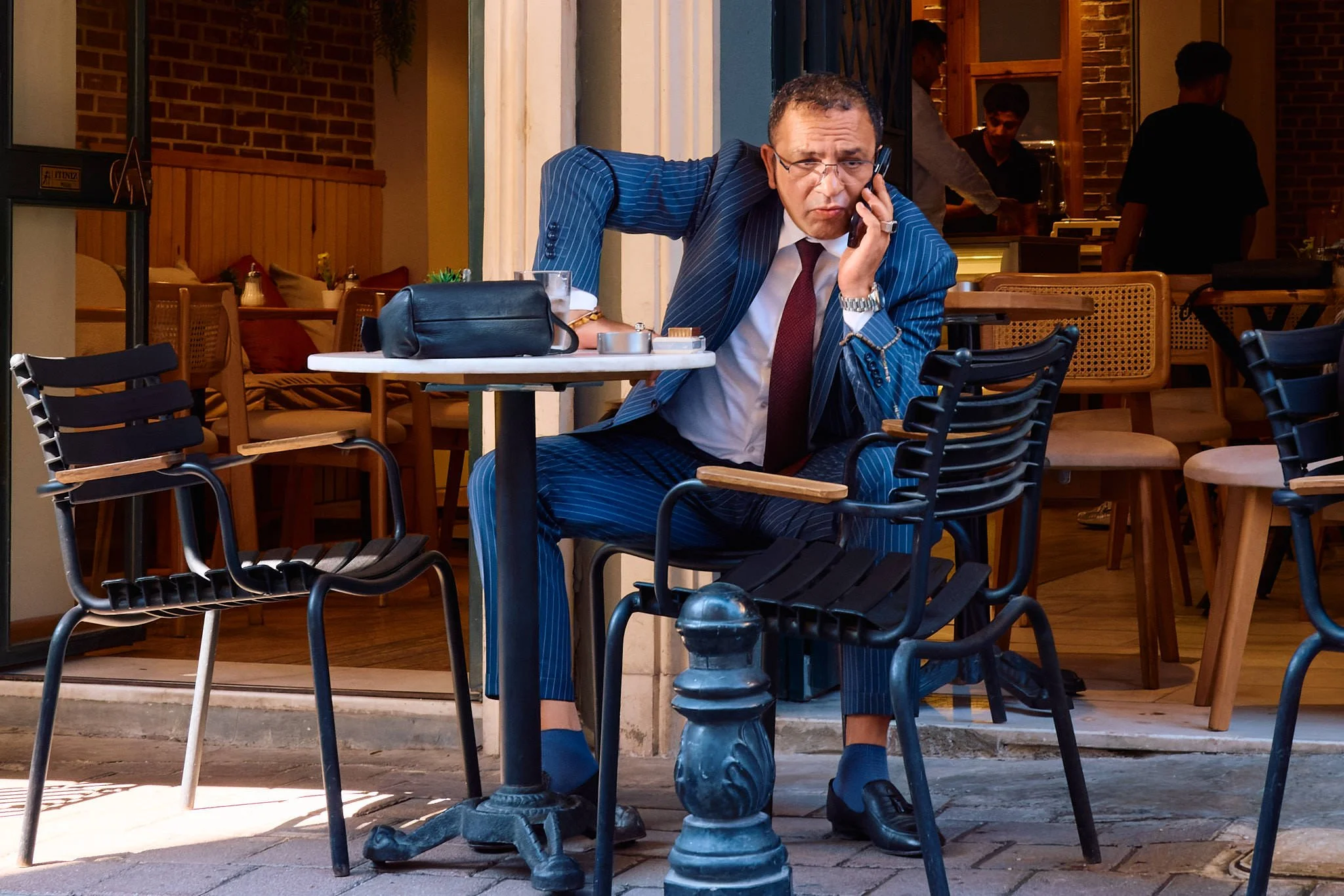 Man in a pinstripe suit and burgundy tie on the phone at a café table in Balat, Istanbul, resting his head on his hand