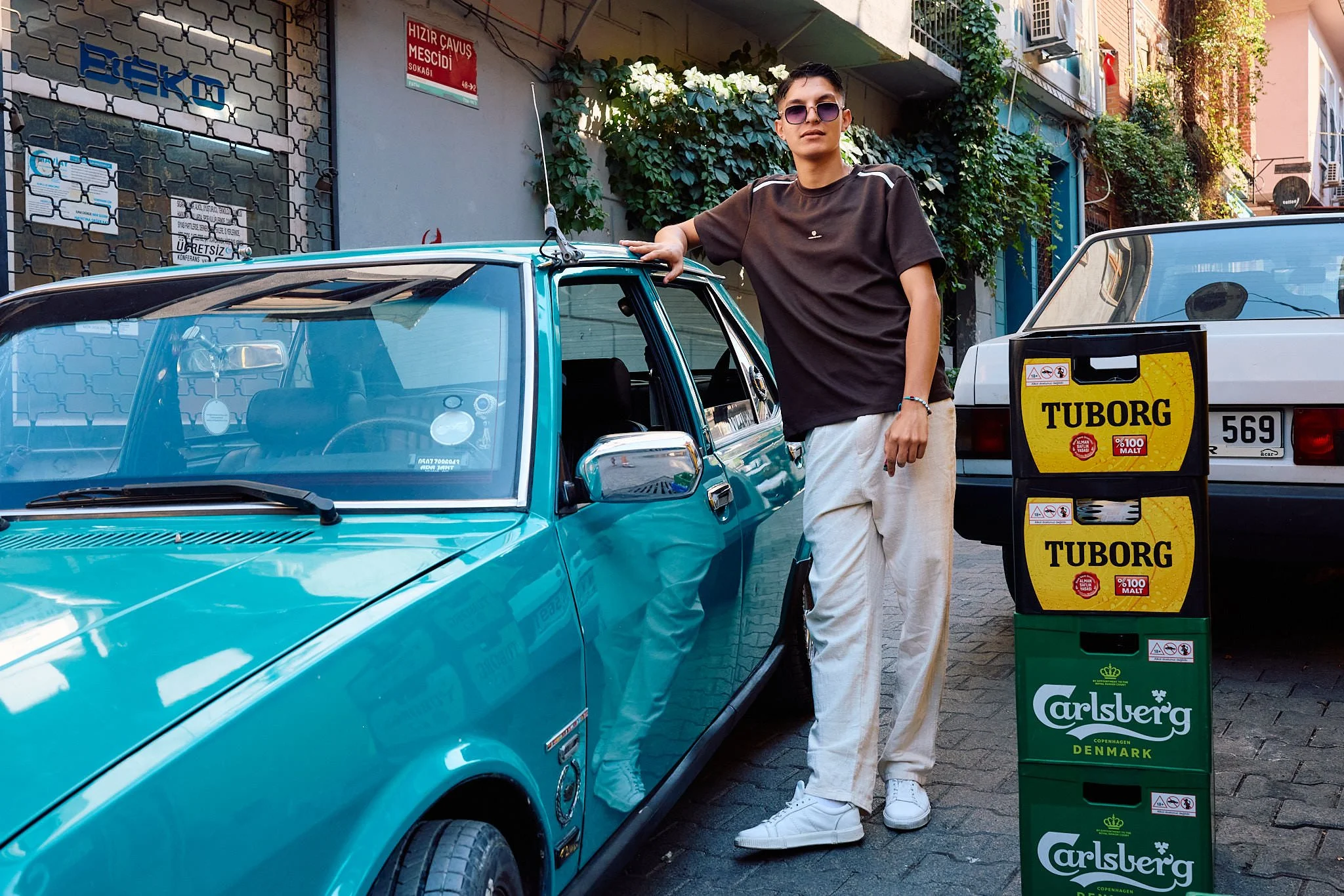 Young man in sunglasses leaning on his polished turquoise vintage car in Balat, Istanbul, beside stacked beer crates