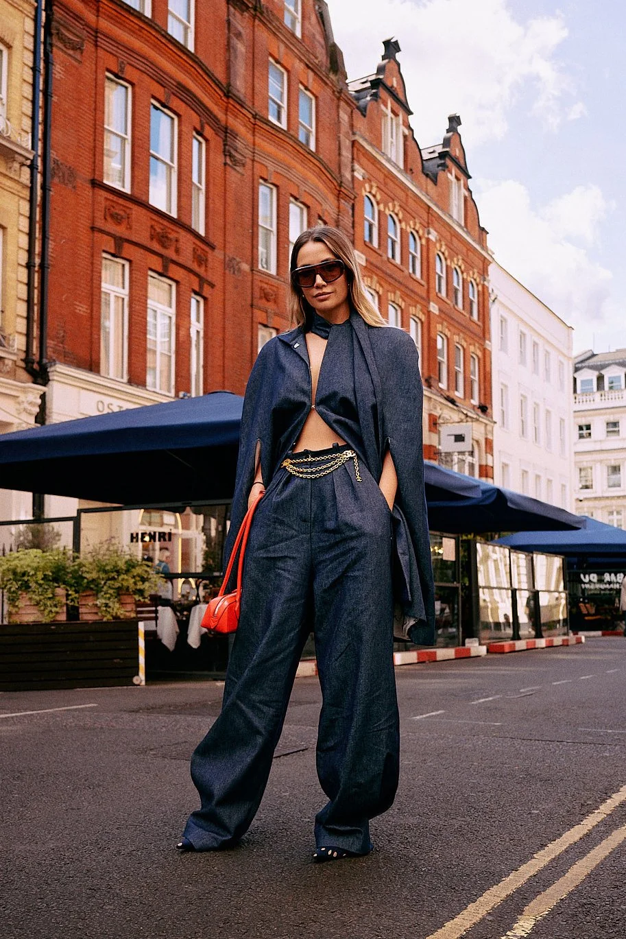 Street fashion portrait in London of woman wearing denim outfit and cape in Covent Garden, glasses, red purse, and red brick building background