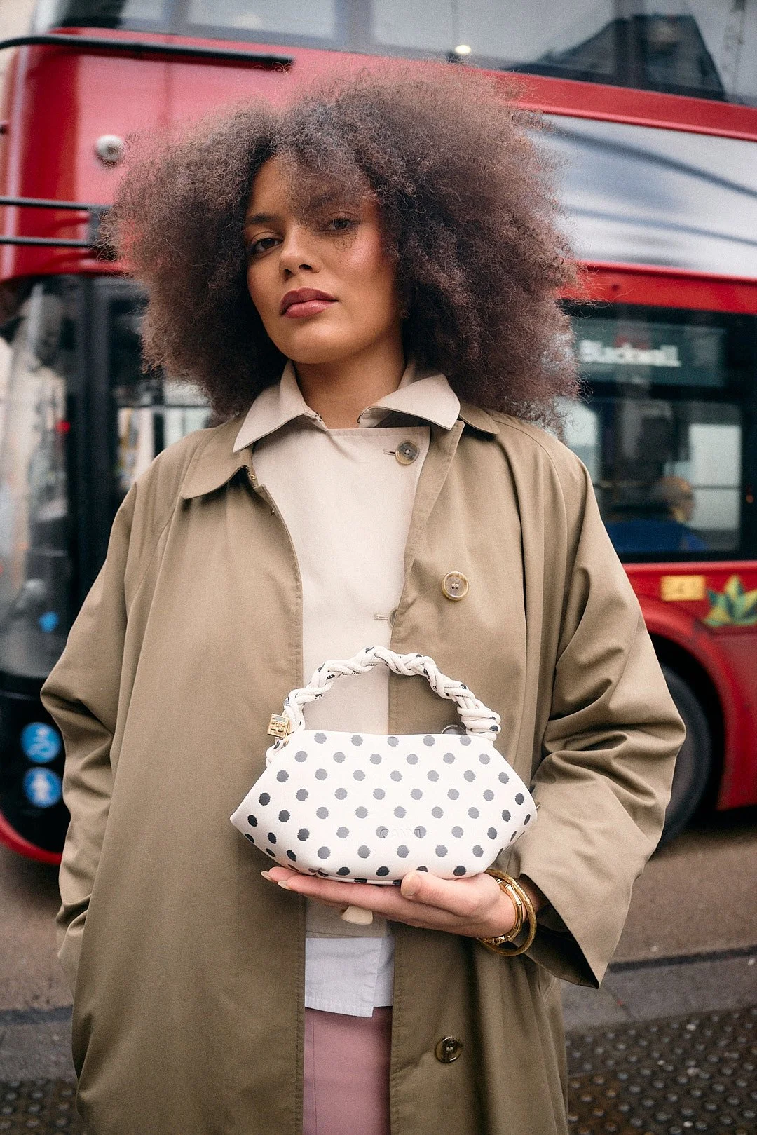Street style portrait at London Fashion Week of woman with curl hair, tan jacket and over coat with polka dot purse held against body with red bus motion blur background