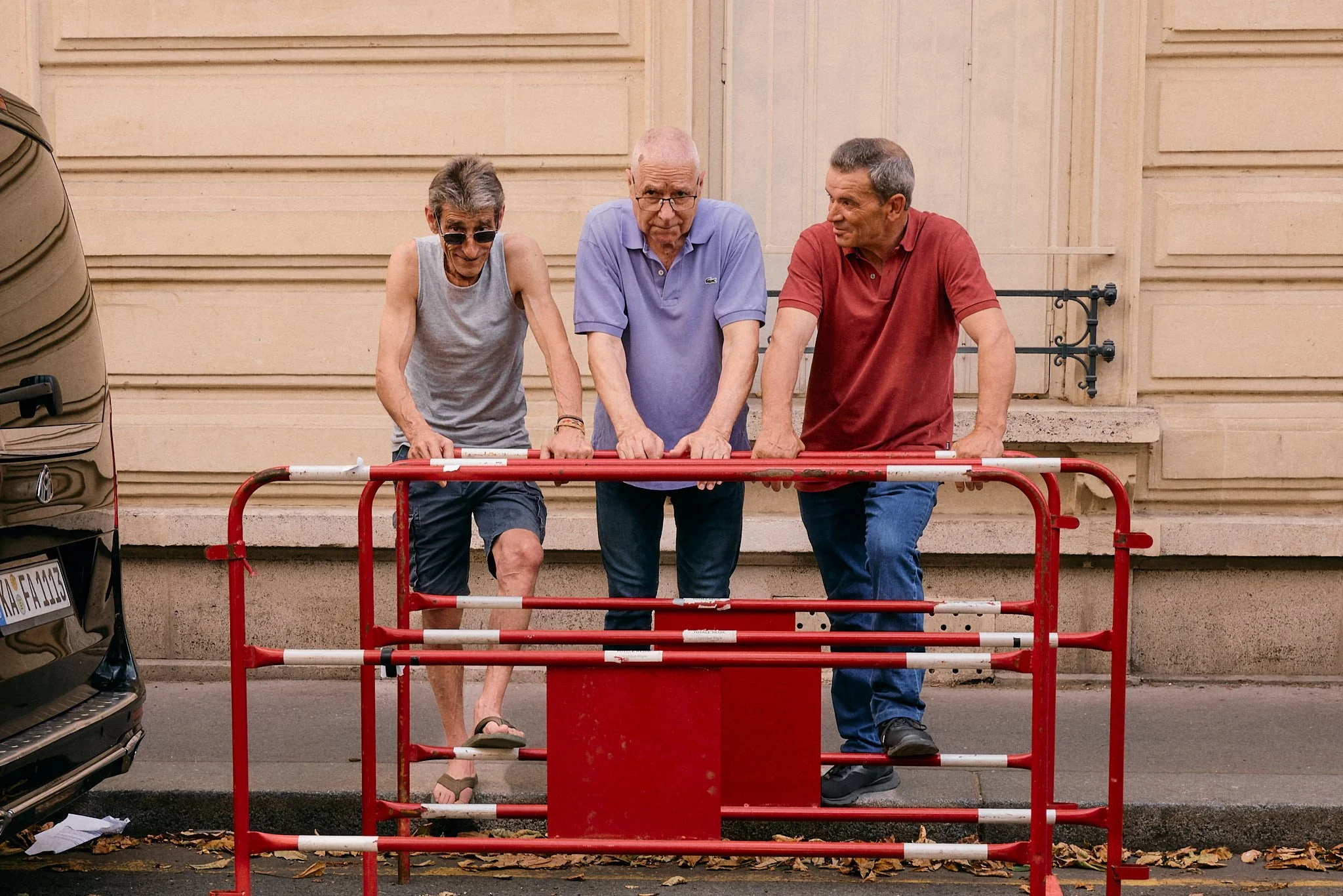 Three older Parisian men in a grey vest, lavender polo and red polo lean on a red barrier outside the Doublet show, Paris Fashion Week SS27 menswear