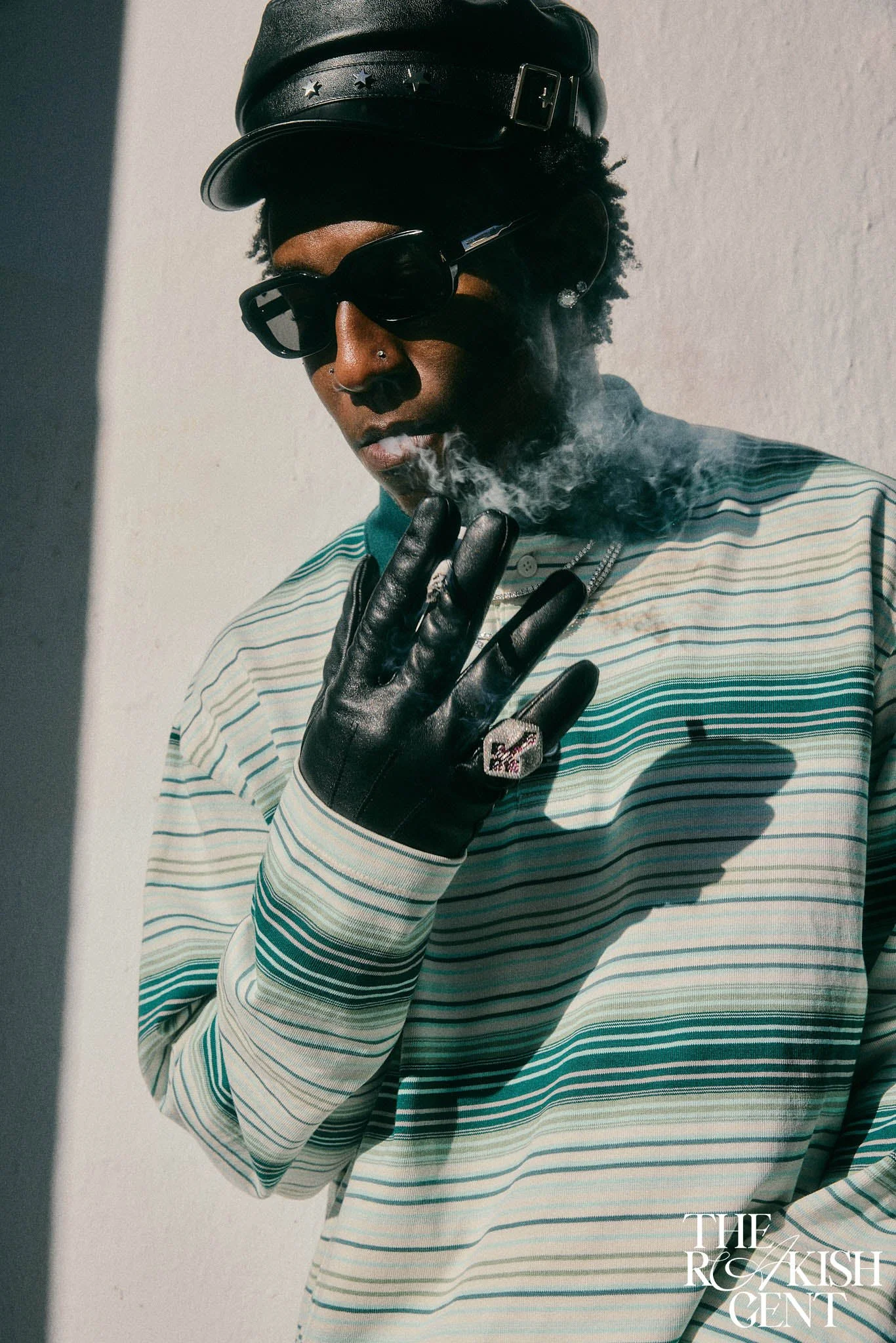 Afrobeats music artist Shallipopi in Paris for The Rakish Gent’s fashion editorial feature, smoking cigarette on wall with striped shirt, leather gloves and hat with sunglasses